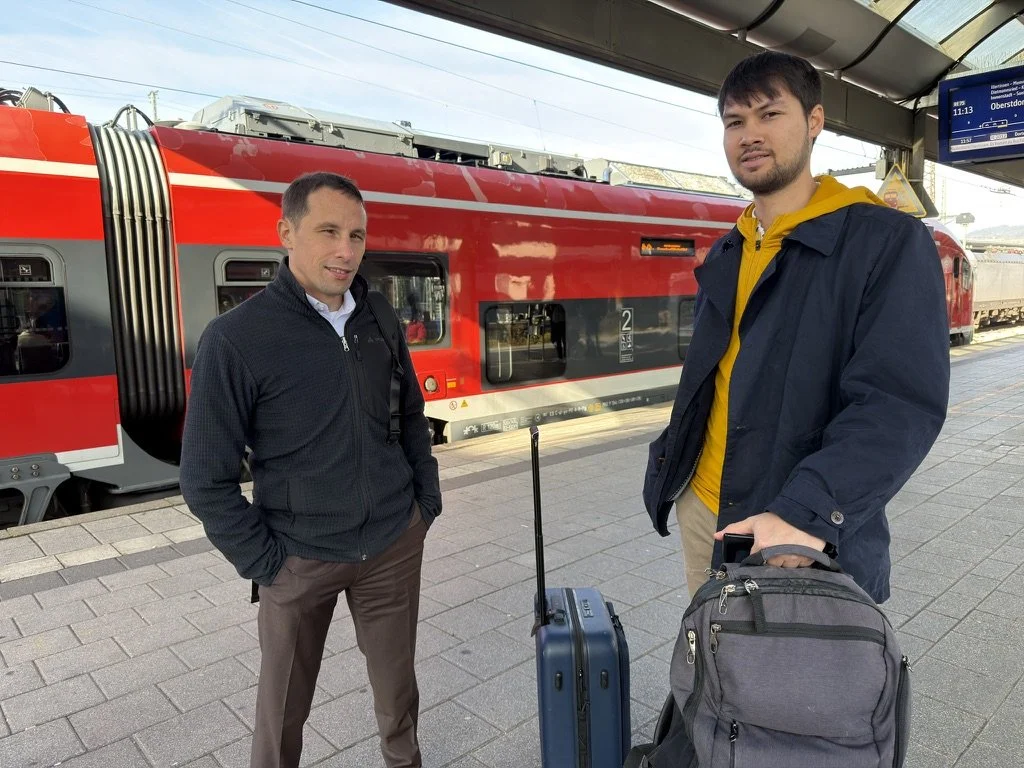 Two men with luggage standing on a train platform in front of a red double-decker train at a station.