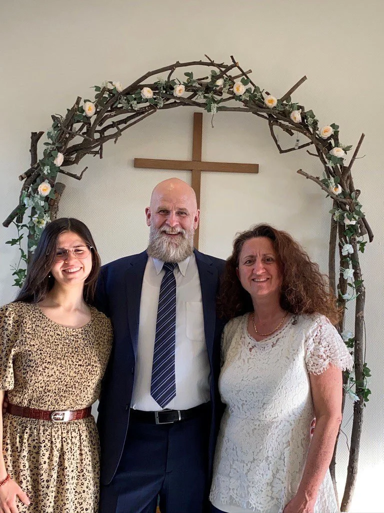 Family of four standing outdoors with trees and a stream, dressed in formal and semi-formal attire, smiling at the camera.