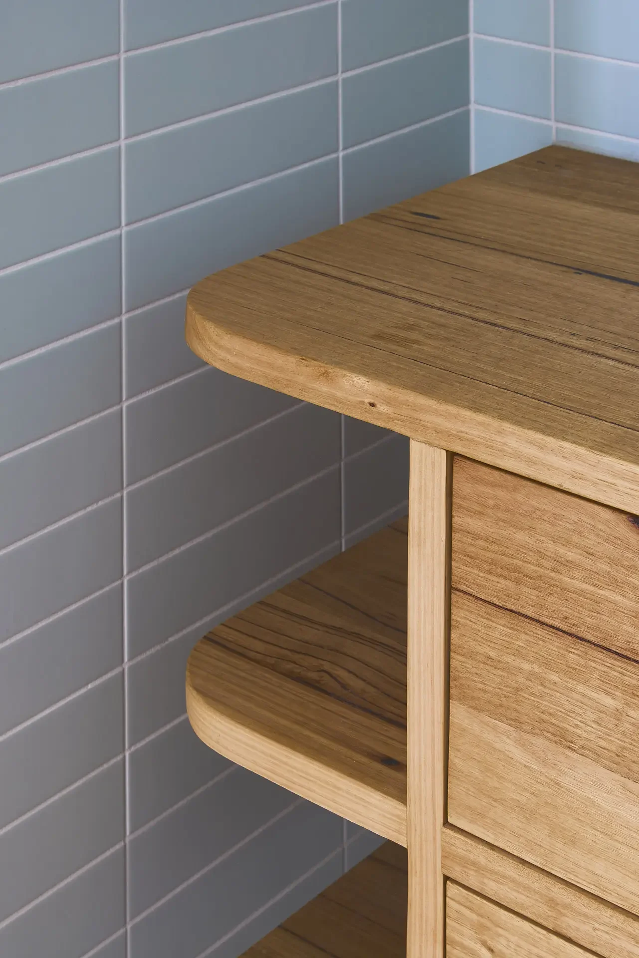 Close-up of a corner in a bathroom with tiled wall, electrical outlet, and a bespoke wooden cabinetry designed and handmade by Anthony at Georgie Wood,