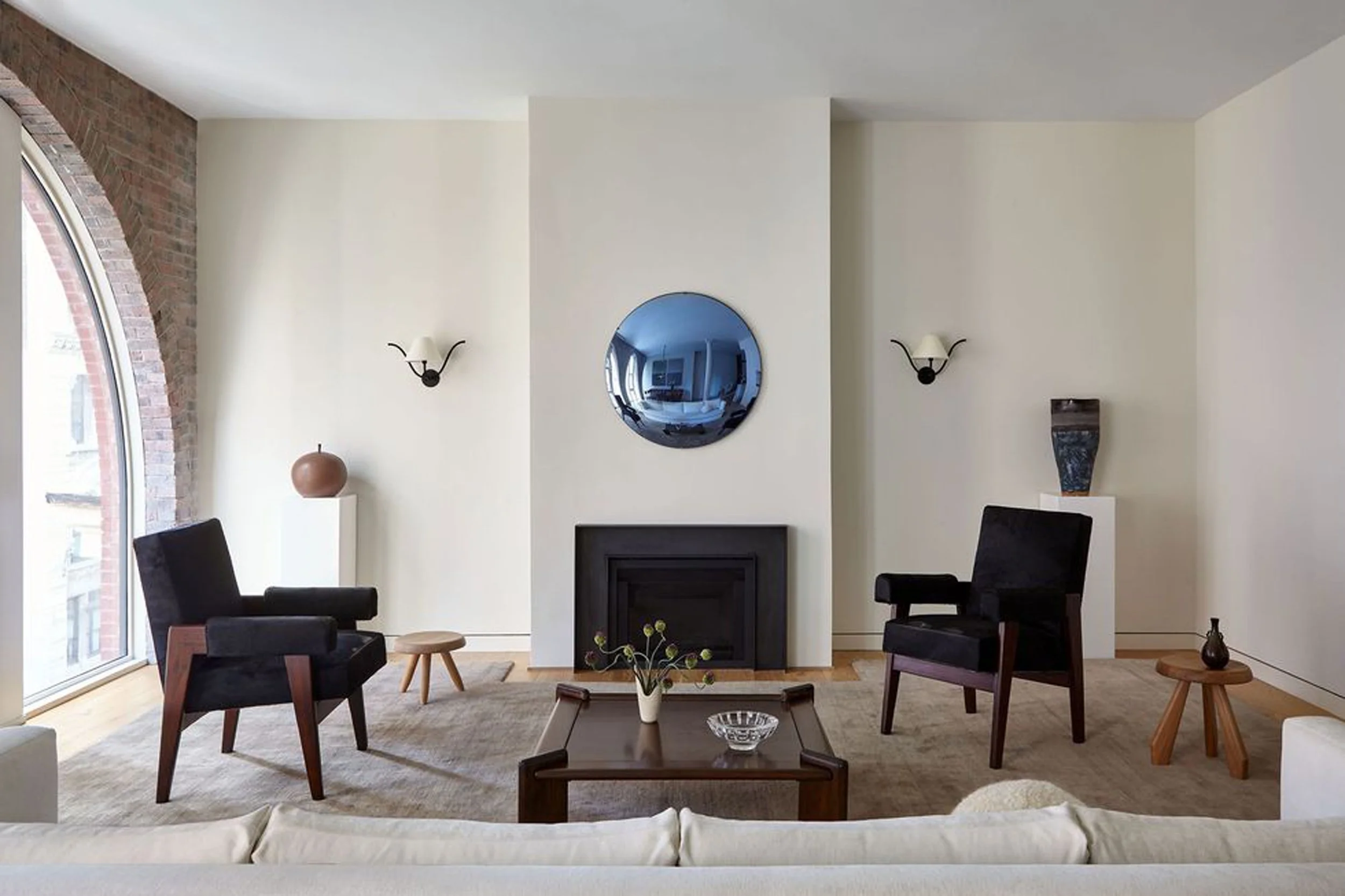 Modern living room featuring a blue convex mirror with two black chairs, wooden coffee table, decorative vase, sconces, arched window, and a minimalist fireplace.