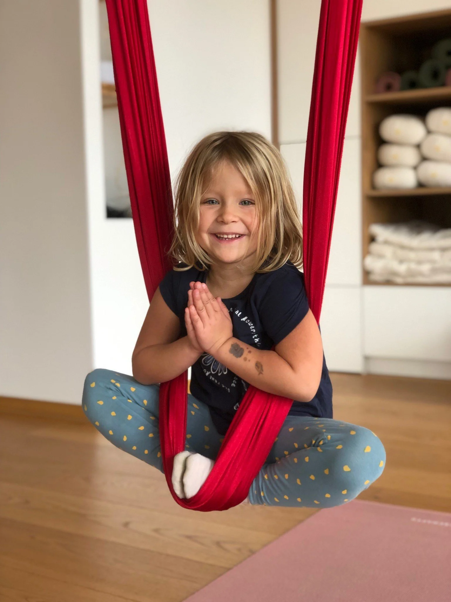 A young girl hanging in a red fabric swing, smiling with hands in a prayer position, wearing blue leggings with yellow spots and a dark T-shirt, indoors with wooden floor and shelves of towels in the background.