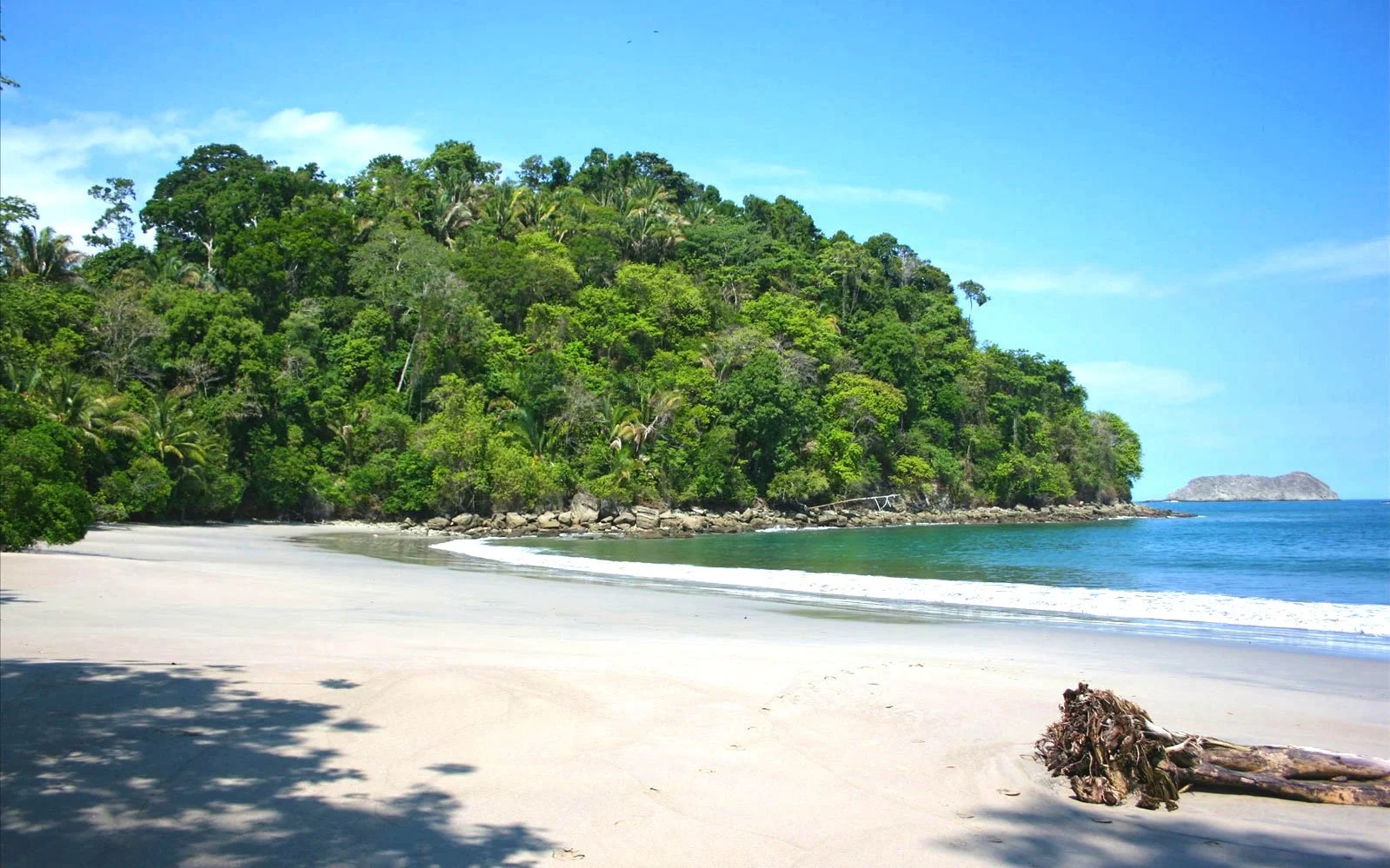 A tropical beach with white sand, lush green trees on a hillside, and a blue ocean with gentle waves, under a partly cloudy sky.