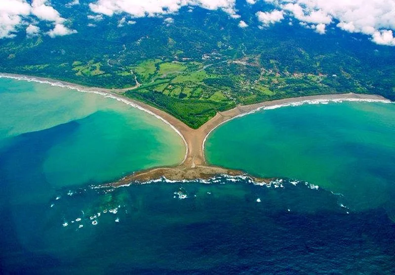 Aerial view of a confluence of two bodies of water separated by a sandy isthmus, with green land and hills in the background.