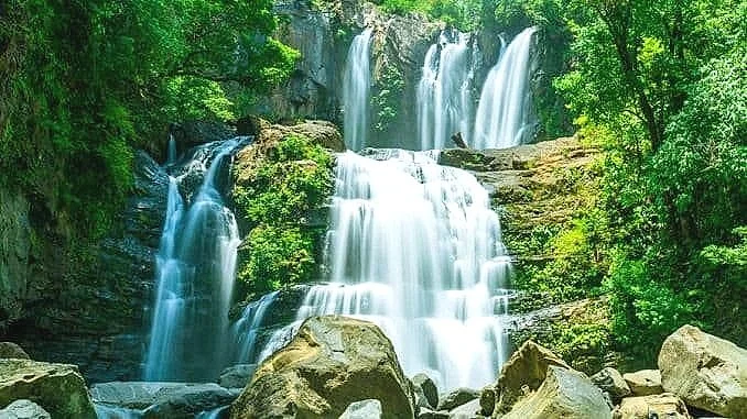 A multi-tiered waterfall flowing over rocks surrounded by lush green trees.