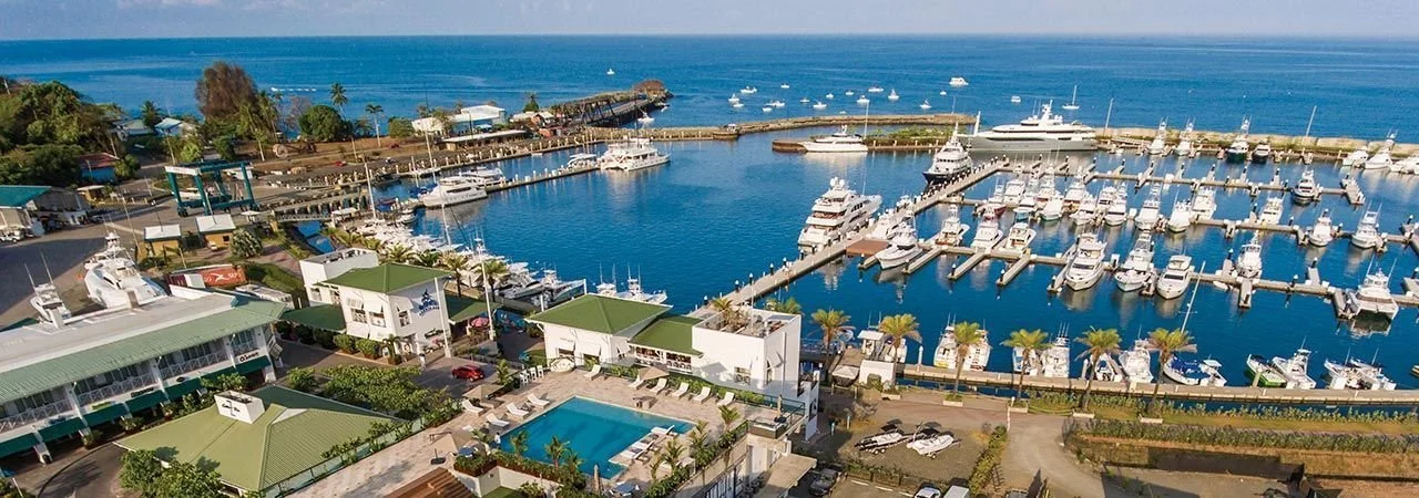 Aerial view of a marina filled with boats and yachts, adjacent to a hotel with a swimming pool, overlooking the ocean on a sunny day.