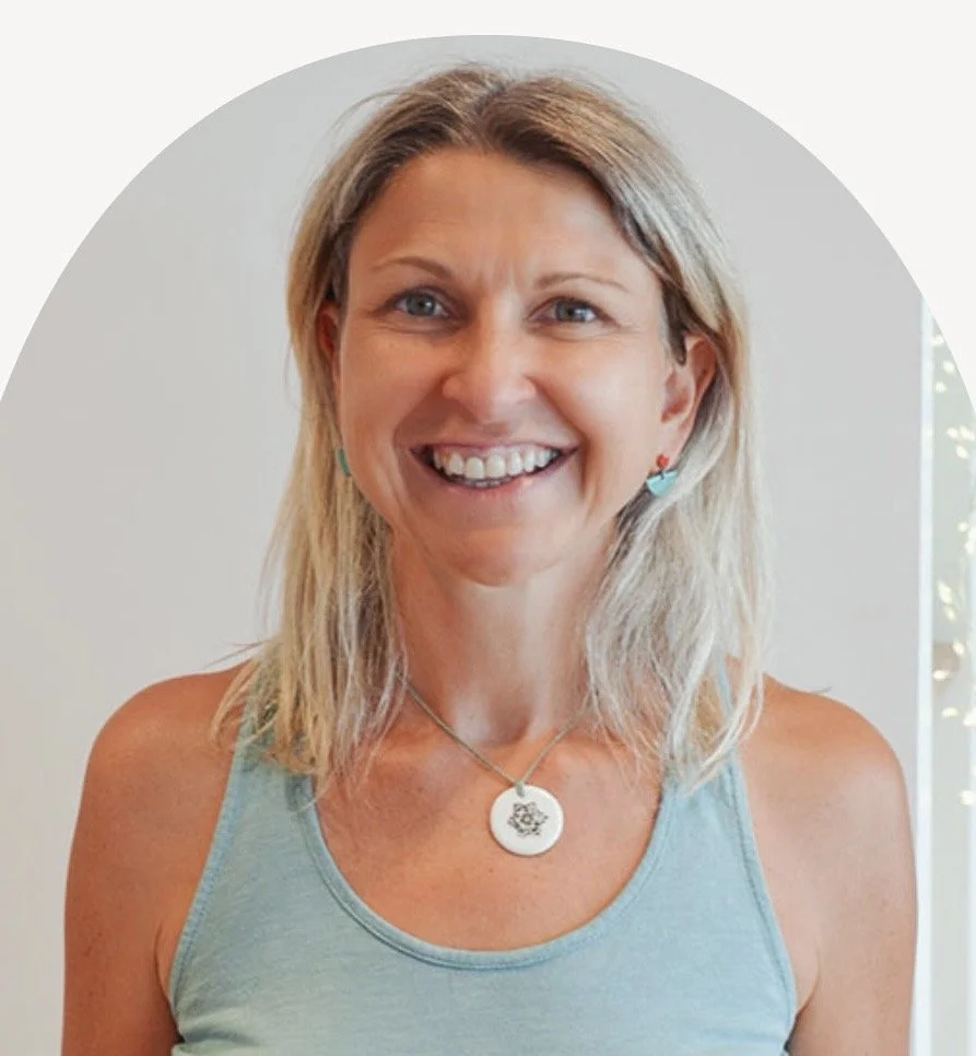 Smiling woman with blonde hair wearing a light blue tank top, necklace, and earrings, standing indoors.