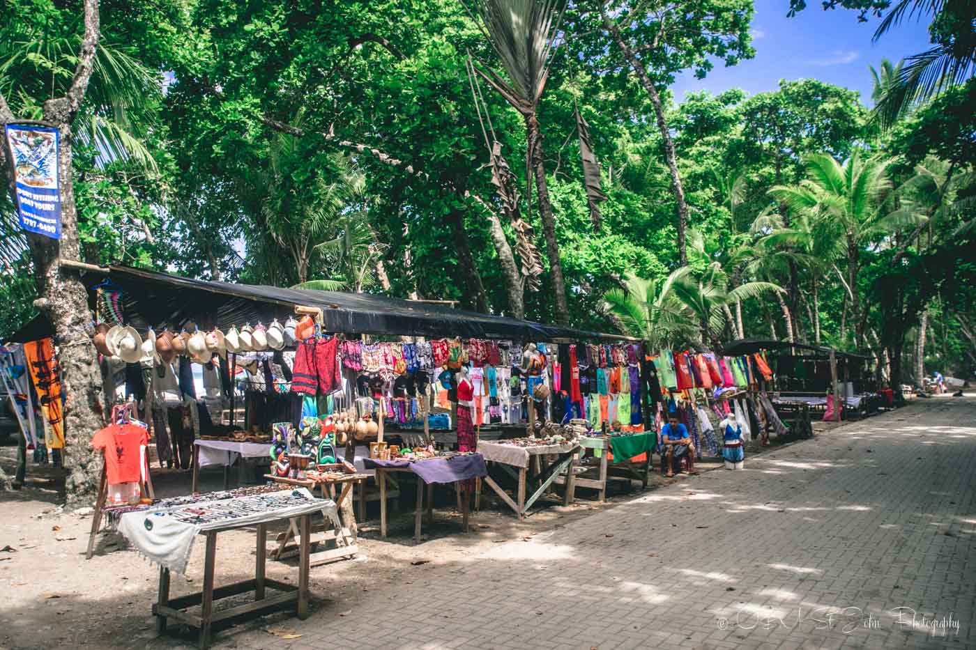 Street market with colorful clothing and accessories under trees on a sunny day.
