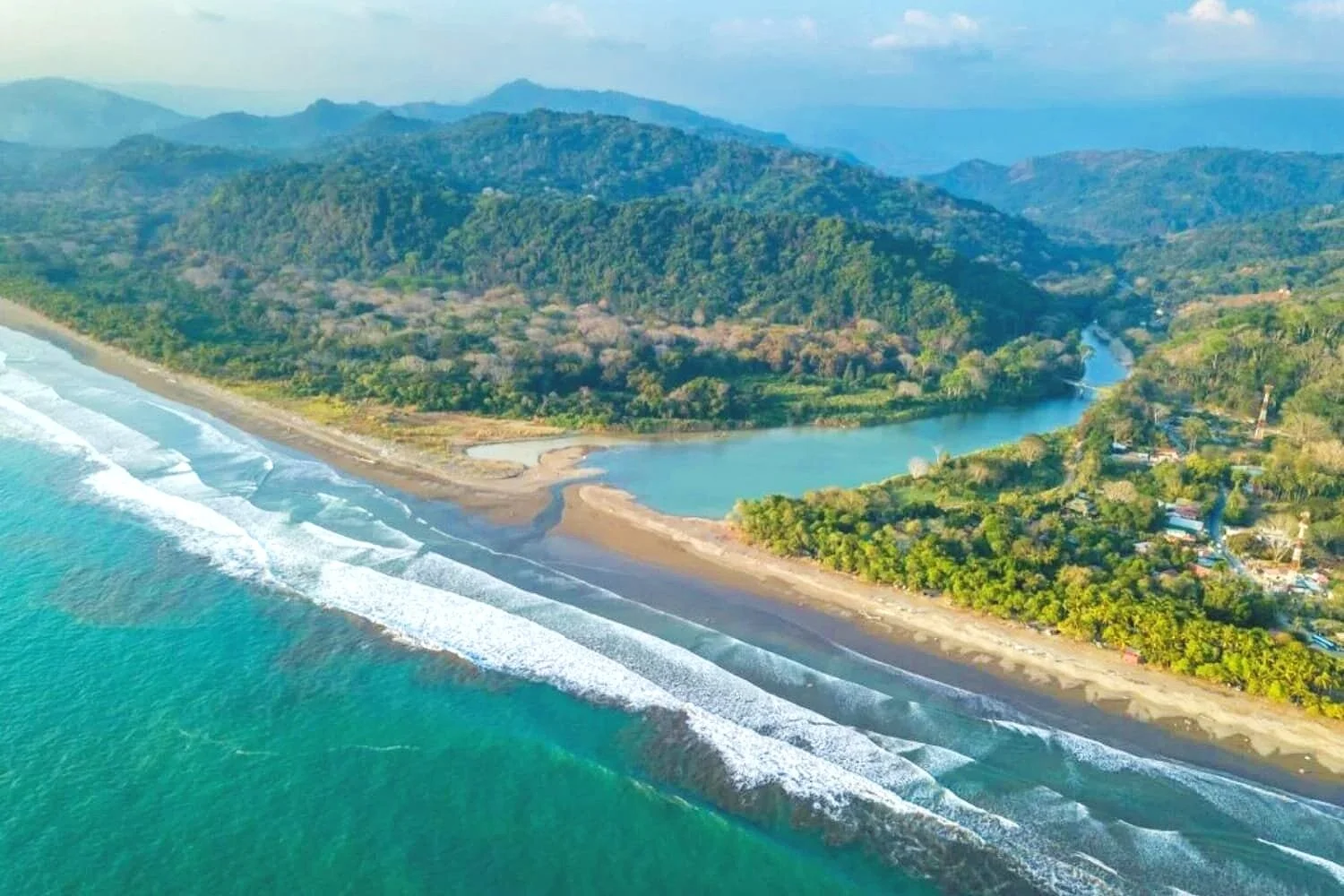 Aerial view of a tropical beach with turquoise water, waves crashing on the shore, surrounded by lush green hills and a river flowing inland from the ocean.