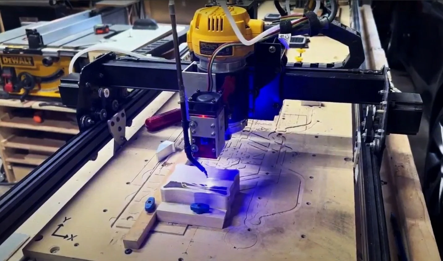 A CNC machine with a robotic arm drilling into a piece of wood with blue laser light in a workshop. Tools and a milling machine are visible in the background.