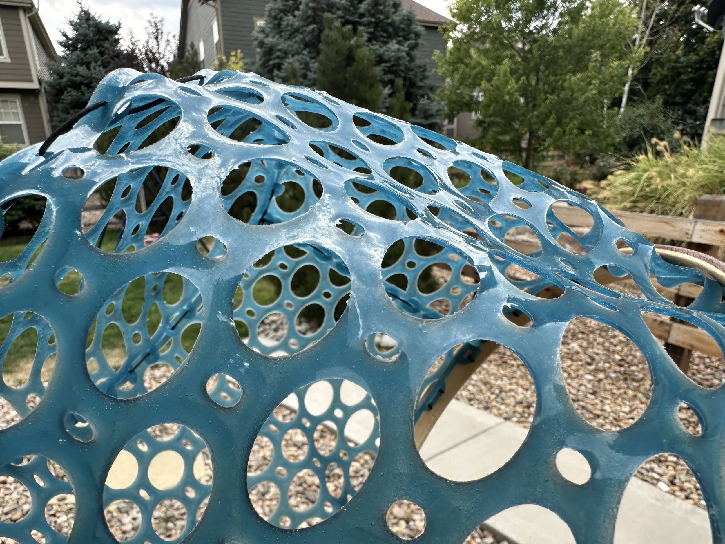 Close-up of a blue decorative outdoor table with circular holes, set in a yard with trees and multi-story houses in the background.