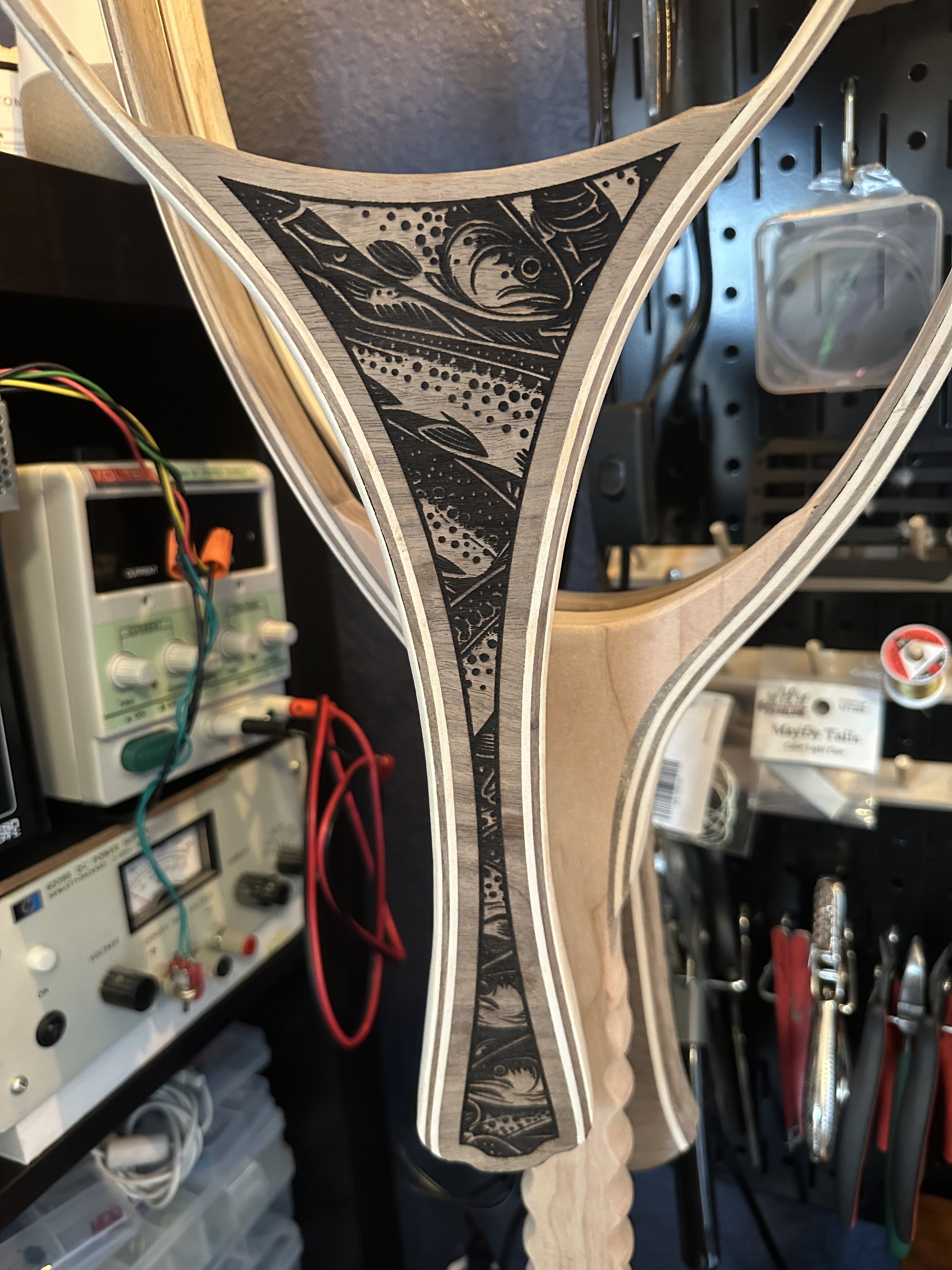A guitar neck with a black and white fish illustration in a triangular shape, mounted on a workbench with electronic testing equipment, wires, and tools in the background.