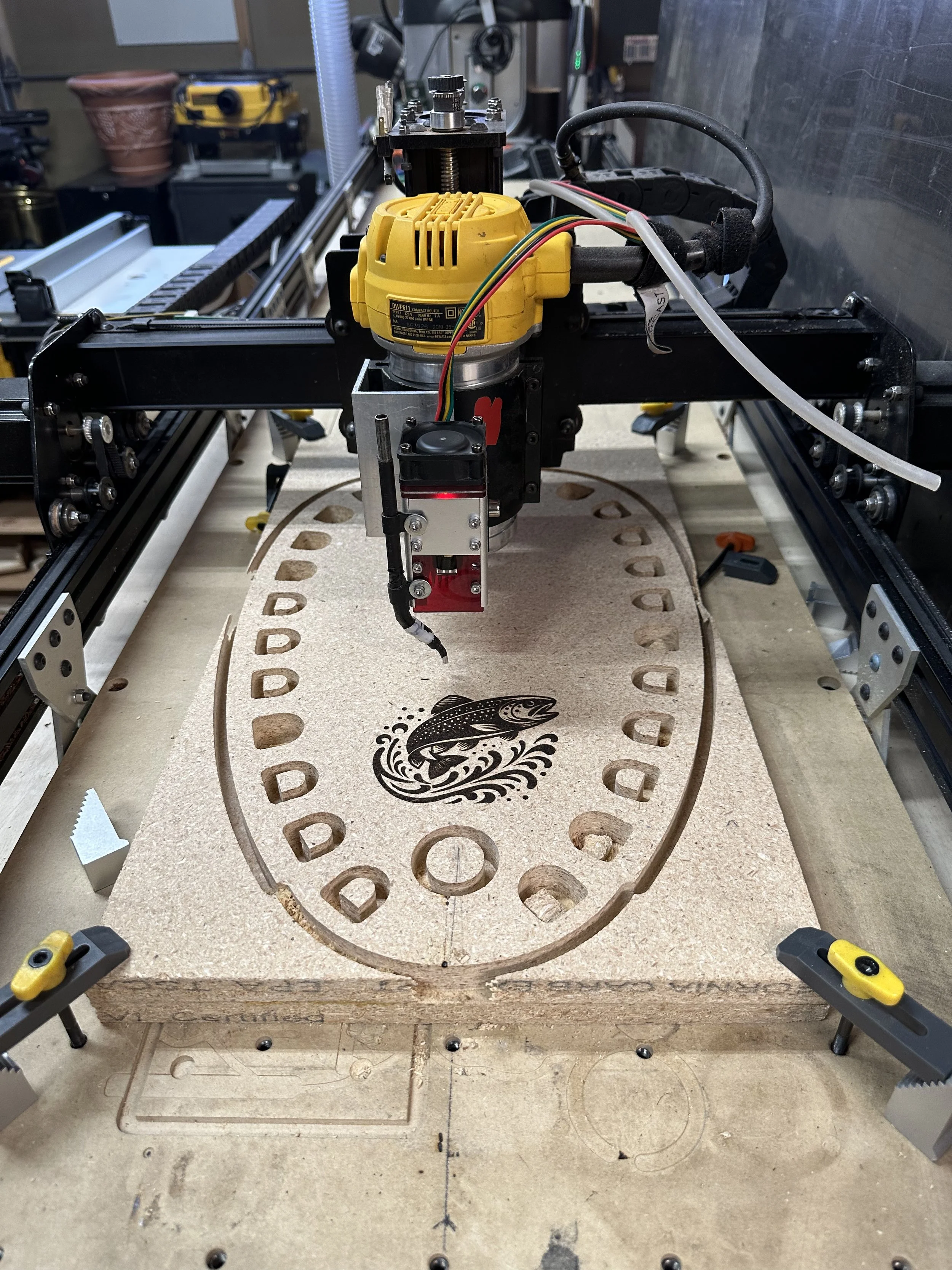 A CNC router machine carving a fish design into a piece of wood with a circular shape, surrounded by tool clamps.