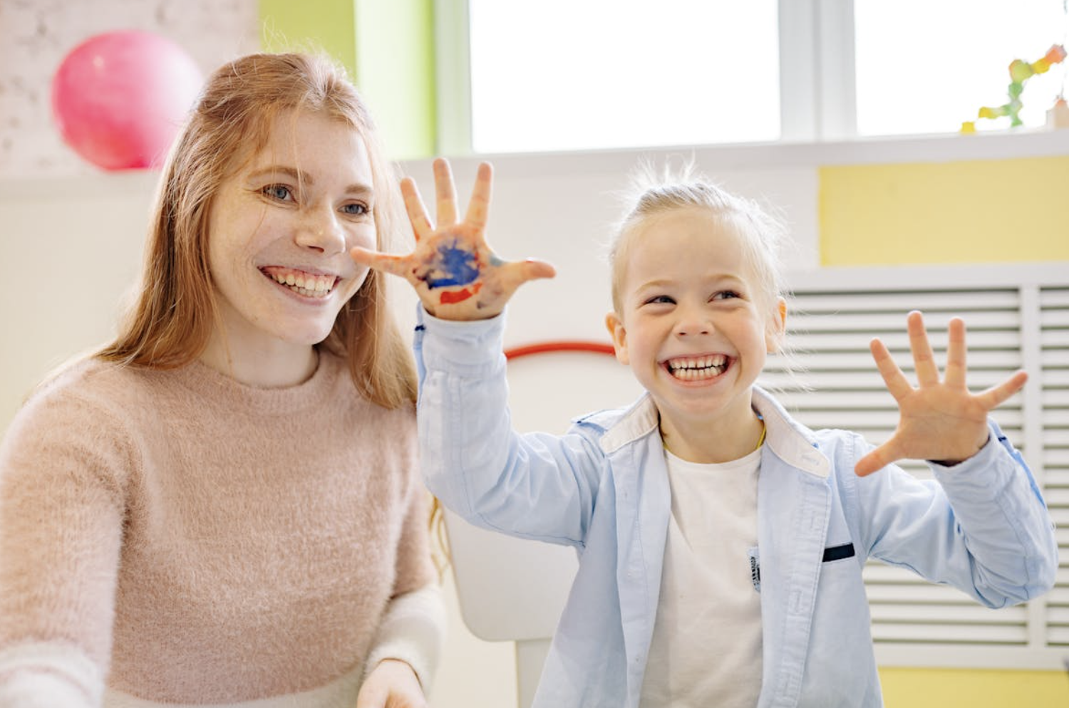 A woman with light skin and red hair smiling next to a young girl with light skin and blonde hair, both showing their painted hands and smiling broadly.
