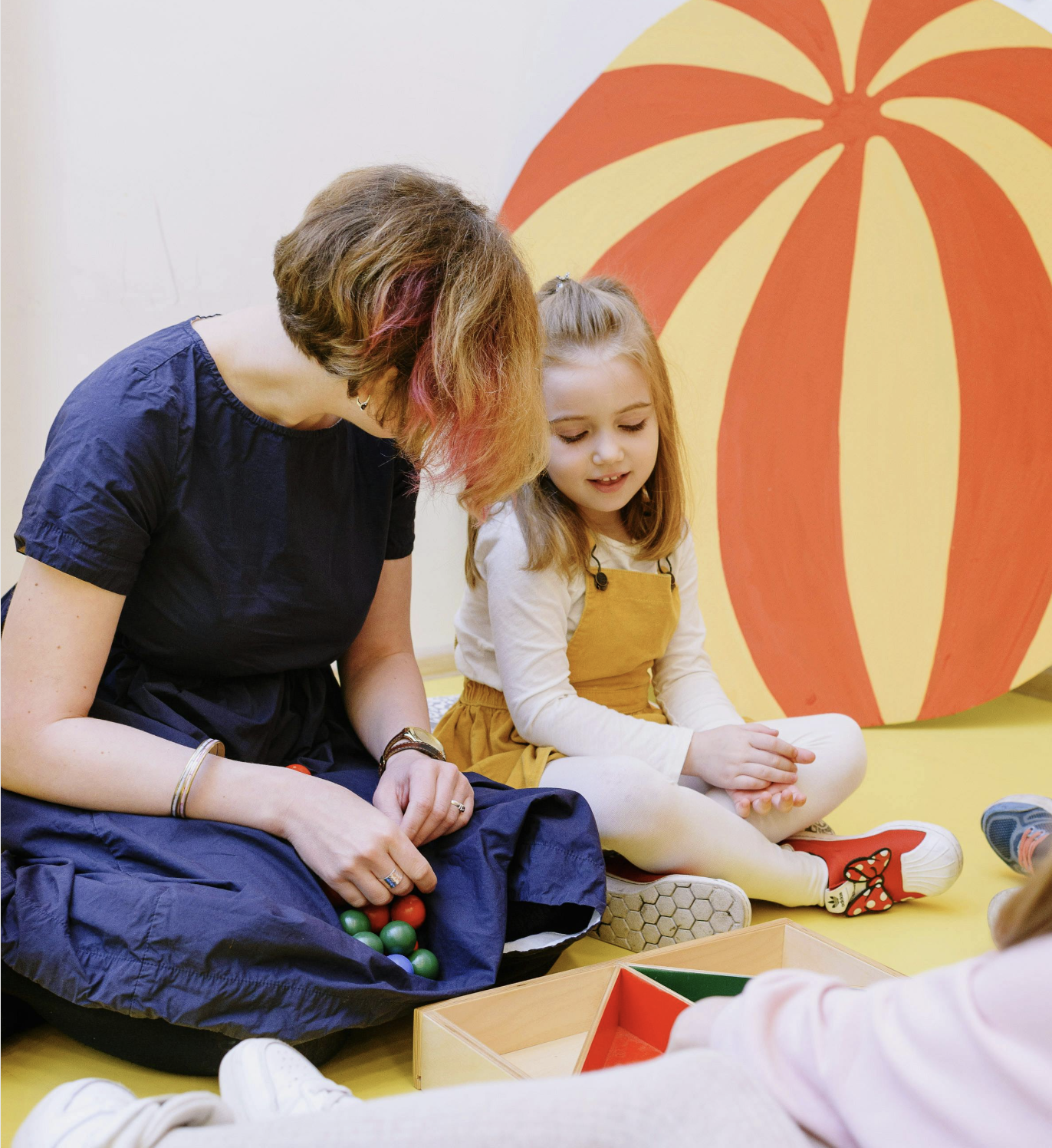 A woman and a young girl sitting on the floor playing a game with colored balls and a wooden container, with a large painted pumpkin in the background.