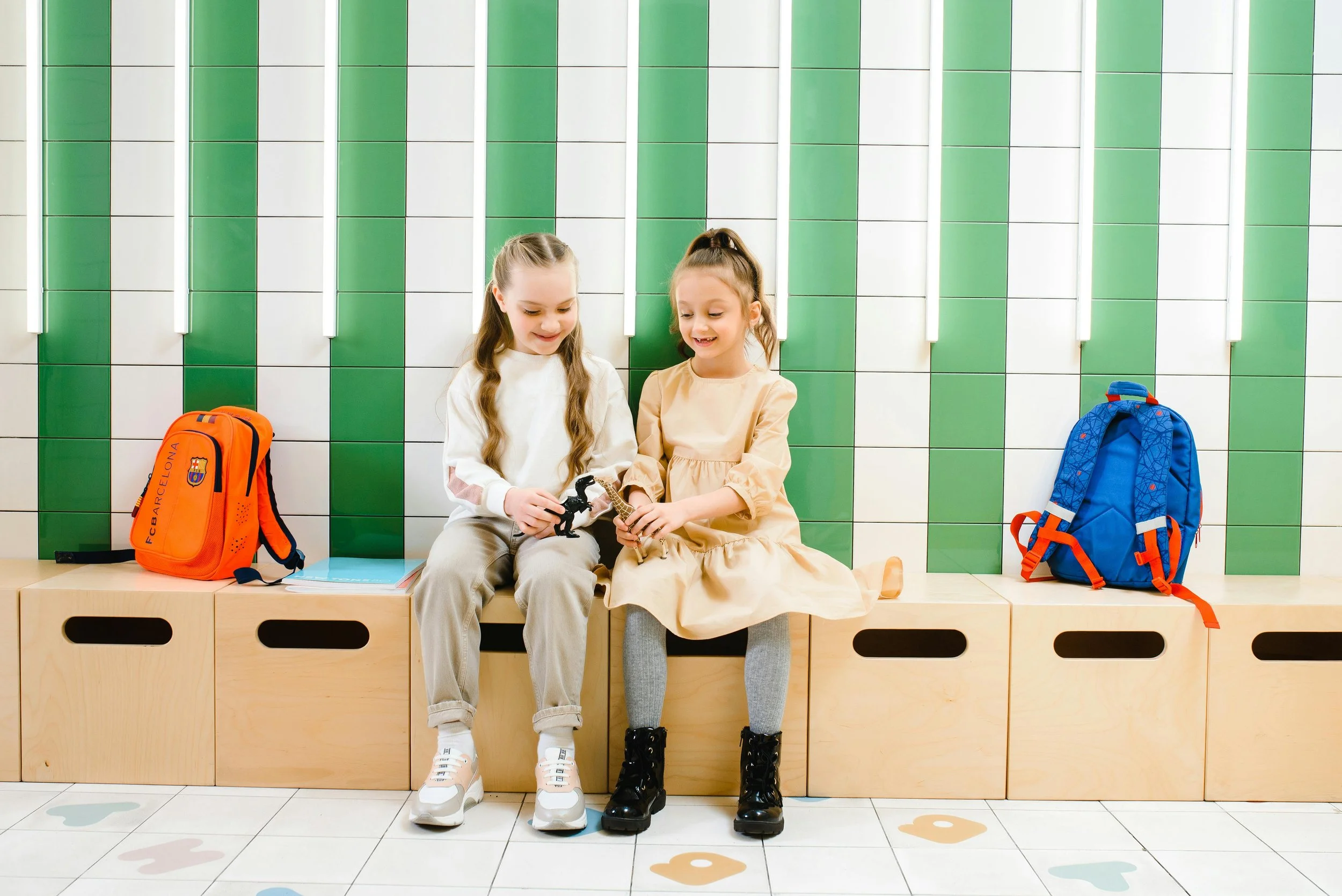 Two young girls sitting on a wooden bench in a school hallway, sharing toys with backpacks on either side, against a green and white tiled wall.