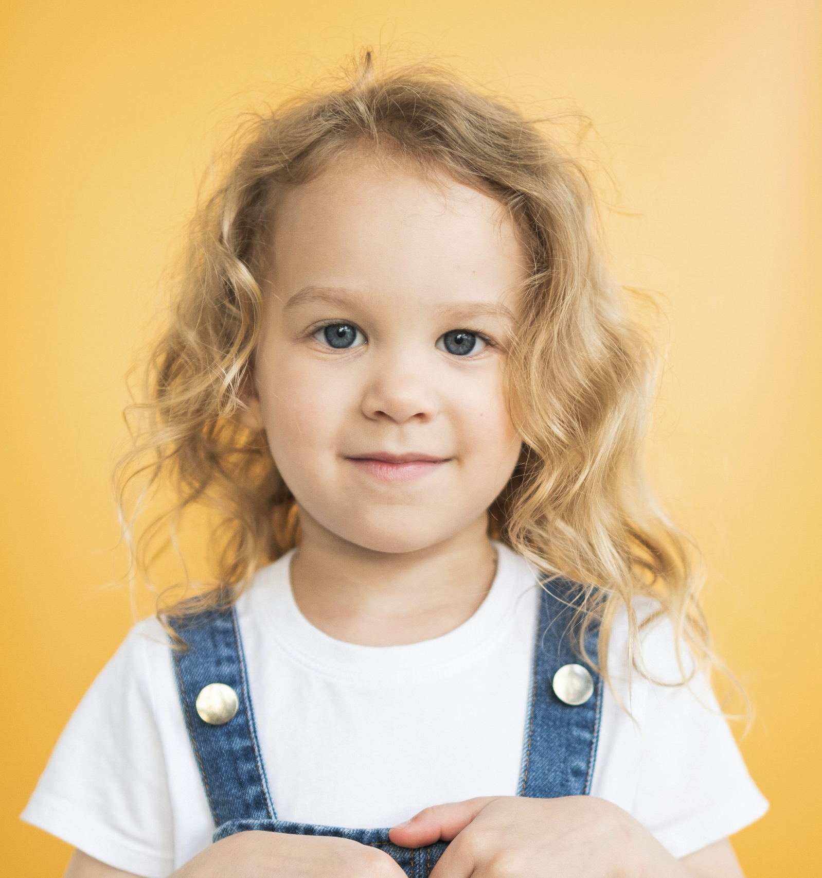 A young girl with curly blonde hair, blue eyes, and a slight smile, wearing a white t-shirt and denim overalls, standing against a yellow background.