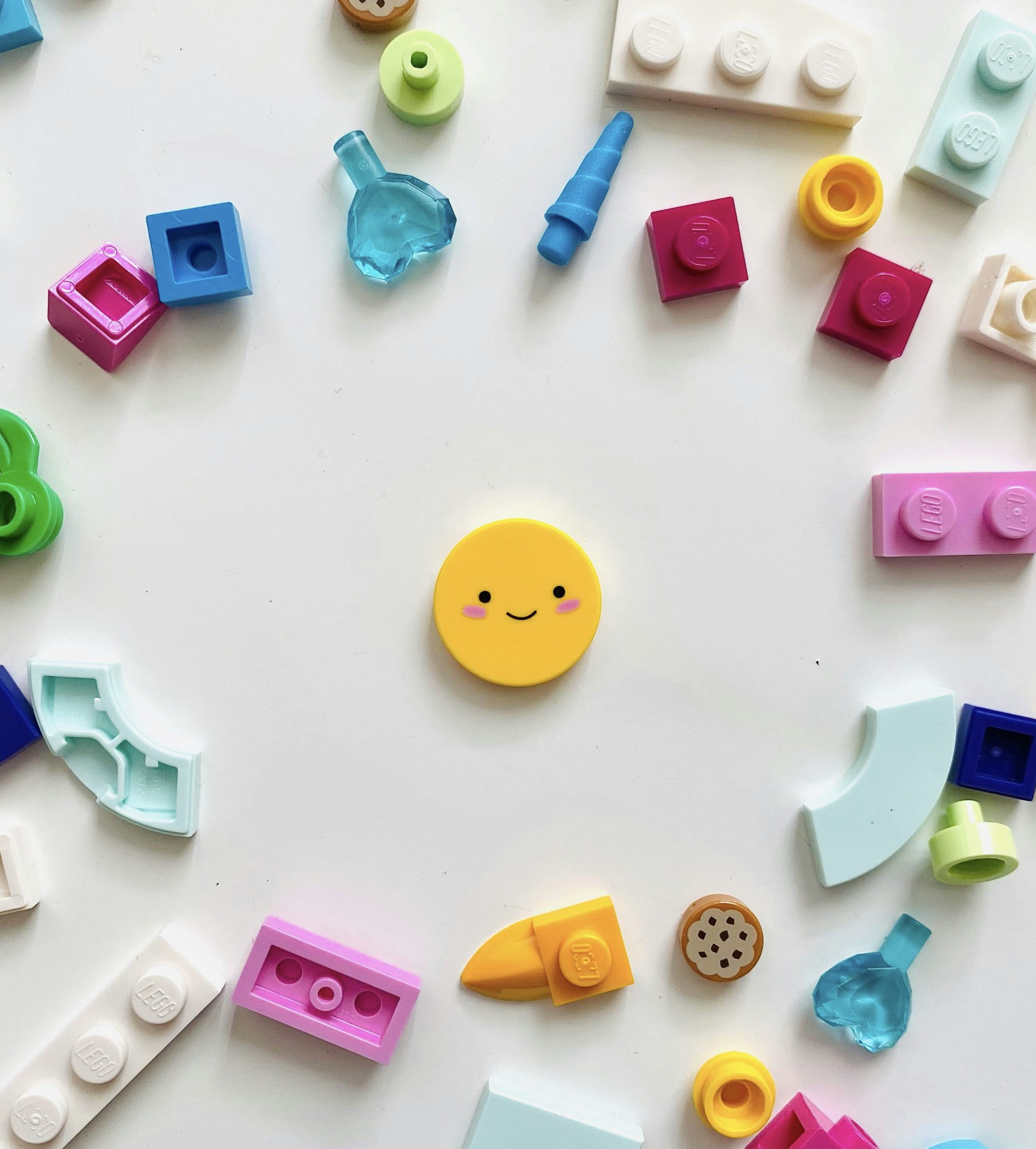 Colorful LEGO pieces arranged in a circle on a white surface, with a smiling yellow round LEGO piece with pink cheeks in the center.