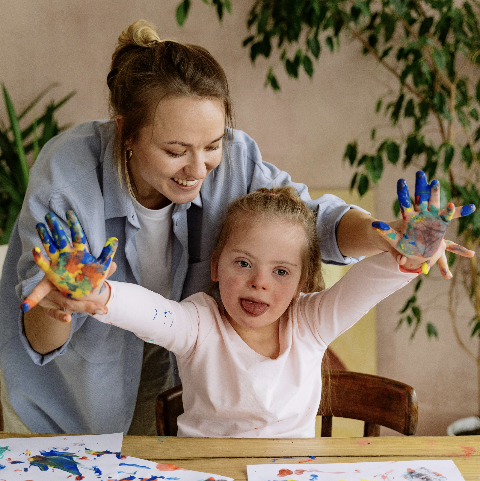 A woman and a young girl with painted hands smiling and having fun together at a table covered with colorful paint and paper.