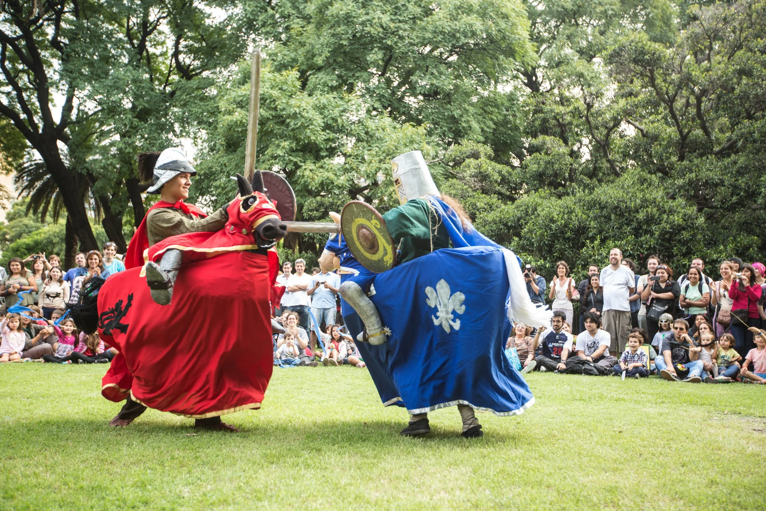 Two people dressed as medieval knights engaging in a mock sword fight during a public reenactment, with a crowd of spectators watching in a park.