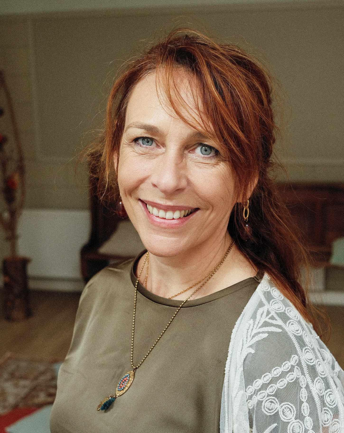 Close-up van een lachende vrouw met rood haar en blauwe ogen, gekleed in een bruin en wit kledingstuk, met sieraden, binnen.