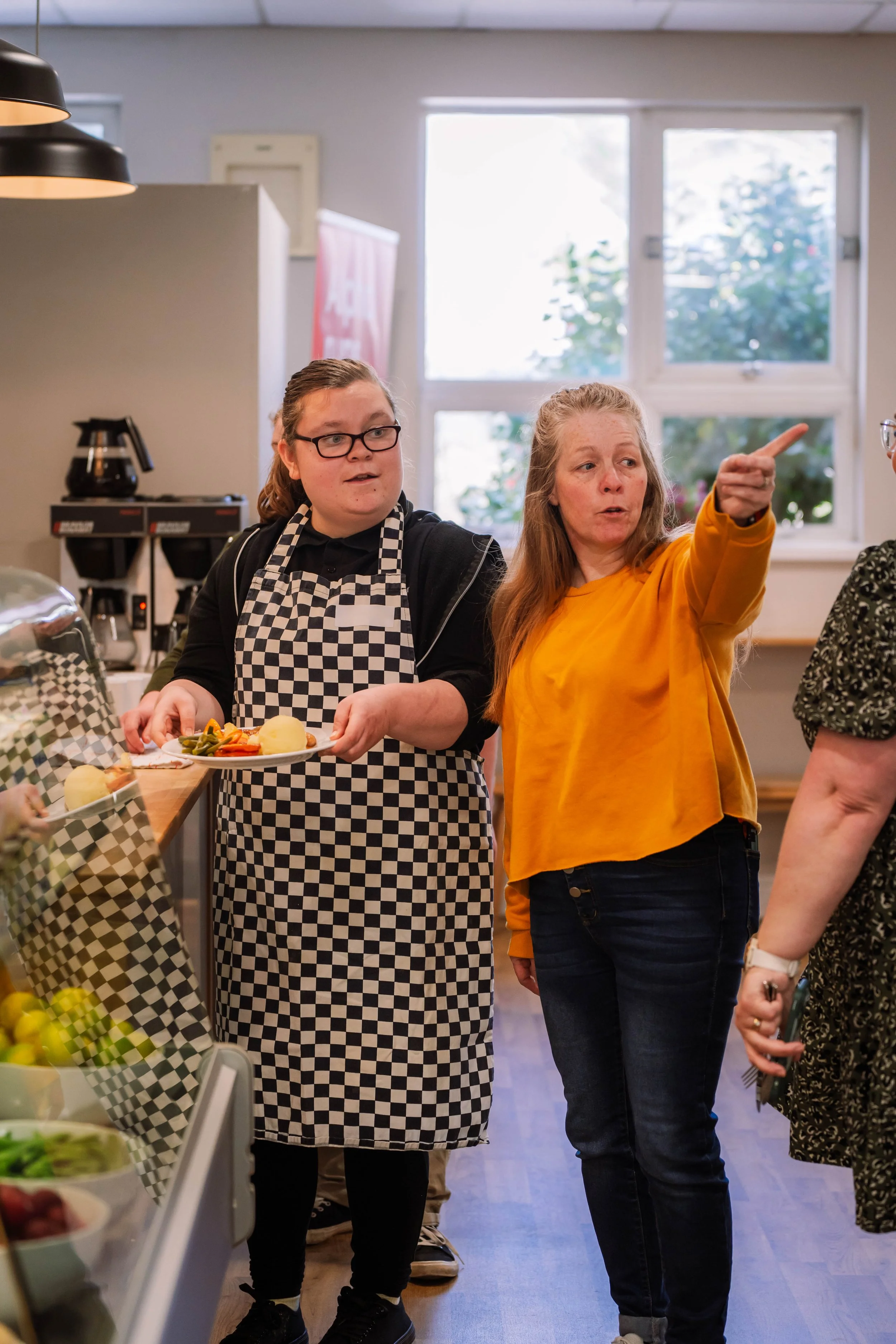 Two women in a cafeteria, one wearing a black and white checkered apron and the other wearing a yellow shirt, are in conversation near a food serving station with fresh fruits, vegetables, and a coffee machine in the background.