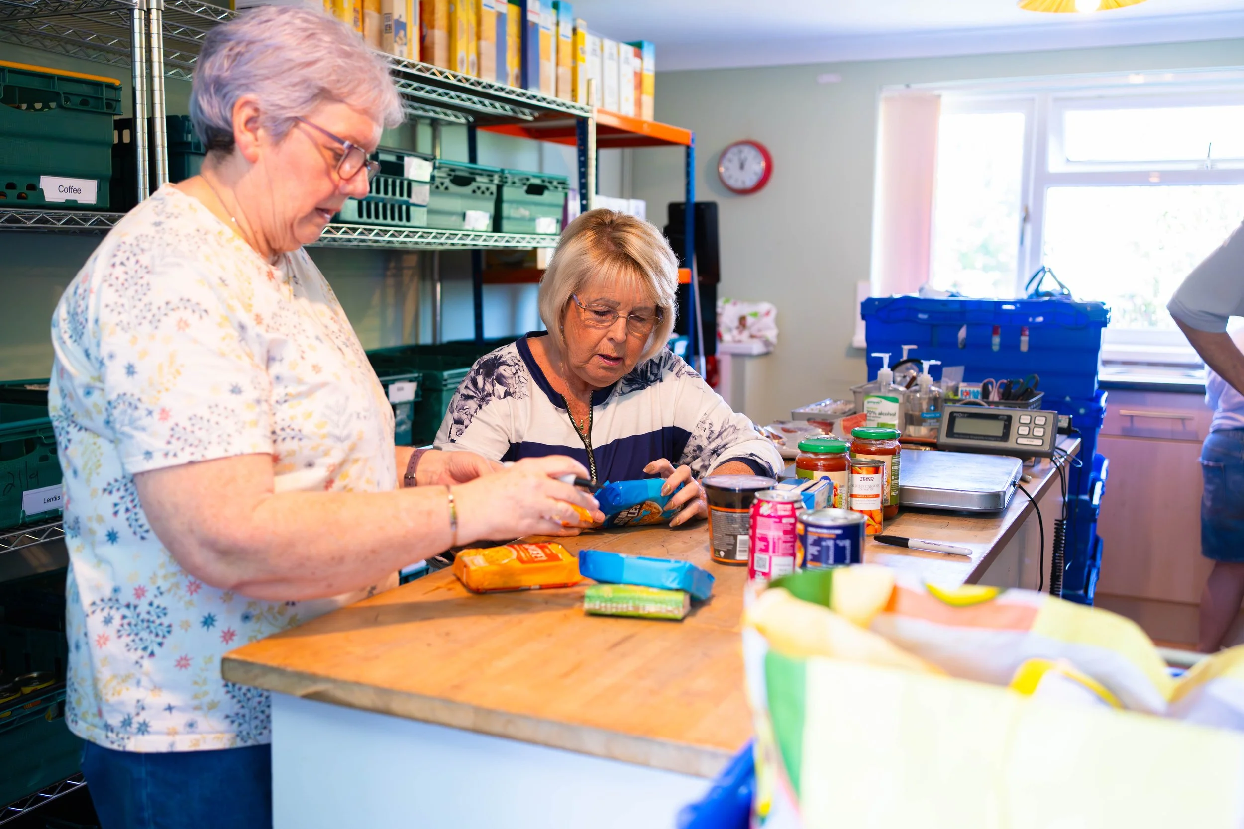 Two elderly women working together in a food pantry, organizing canned goods on a wooden table.