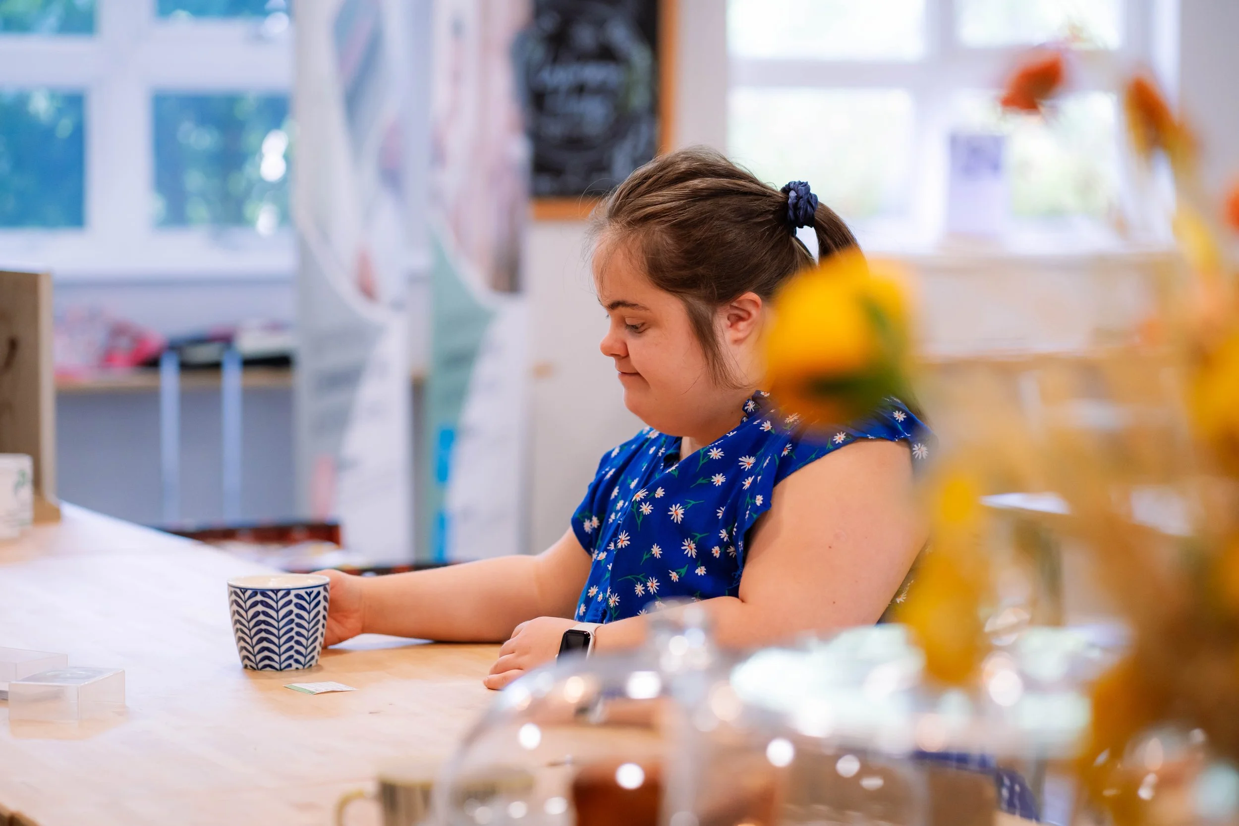 A young girl with dark hair tied in a ponytail, wearing a blue dress with white daisies, sits at a table in a bright room. She is holding a mug with a navy blue pattern and looking down at the table.