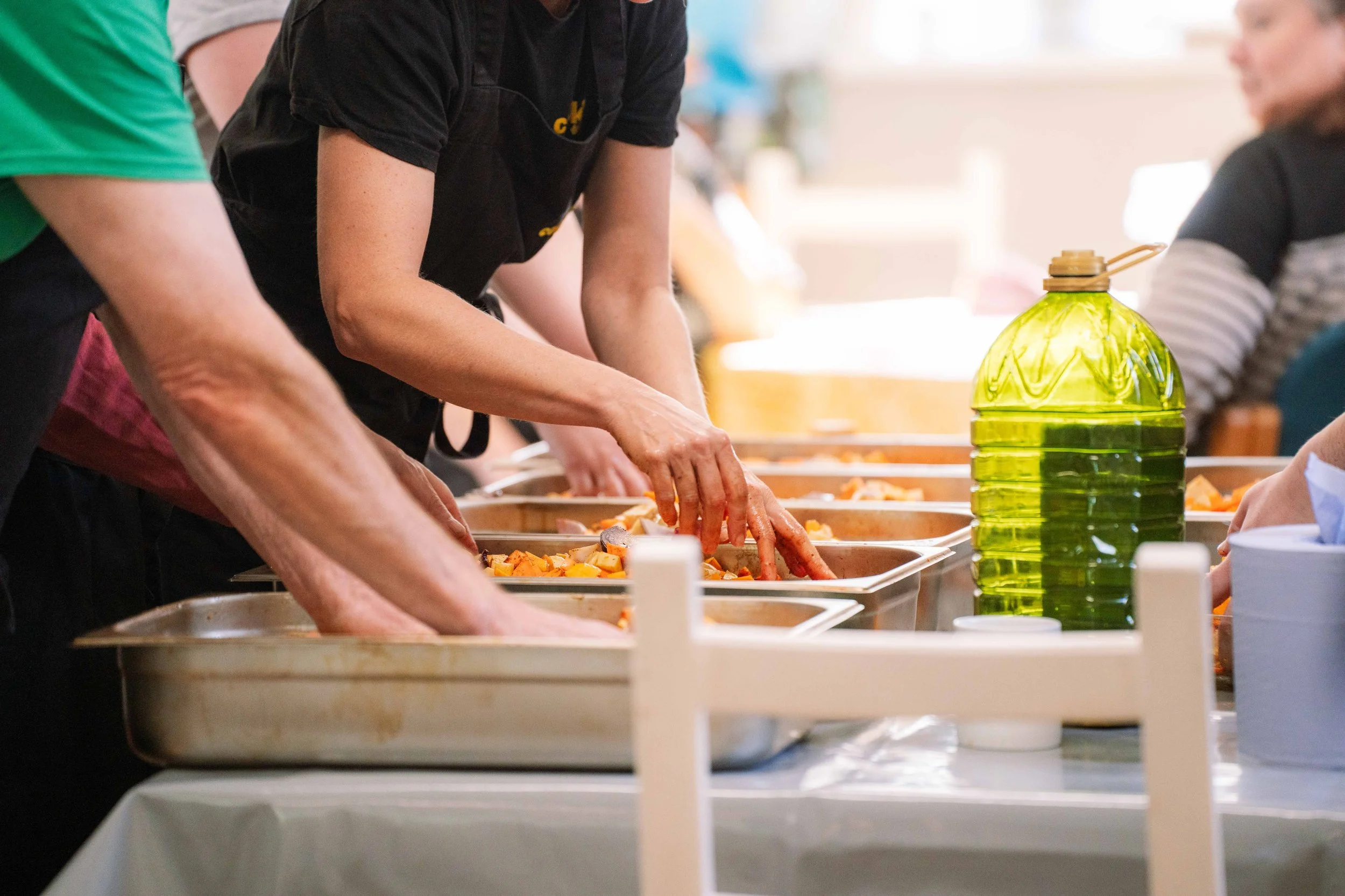 People serving food at a buffet table with trays of cooked vegetables and a yellow plastic water bottle.