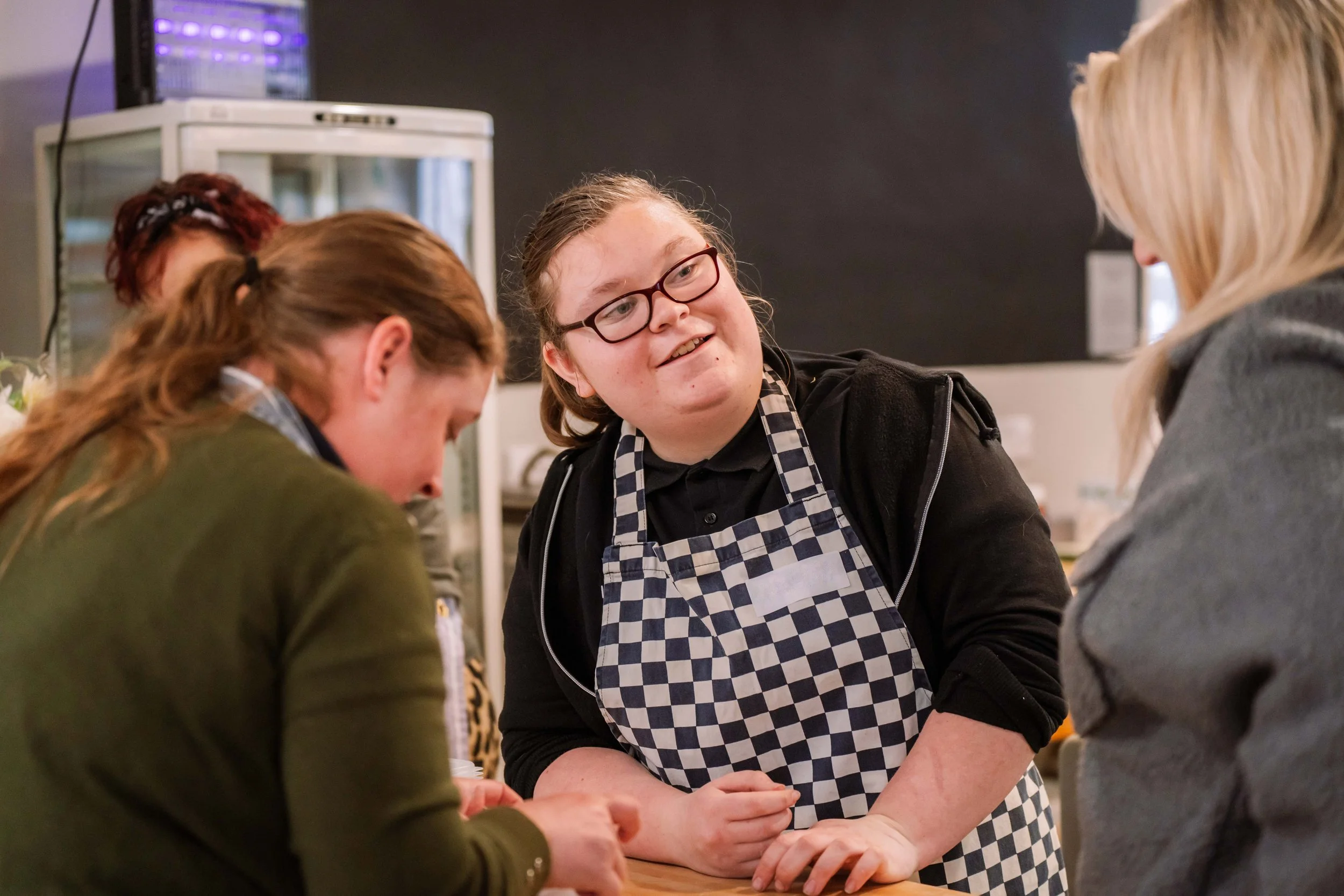 People interacting in a kitchen or cafe setting, with a woman wearing a checkered apron smiling and talking to others.
