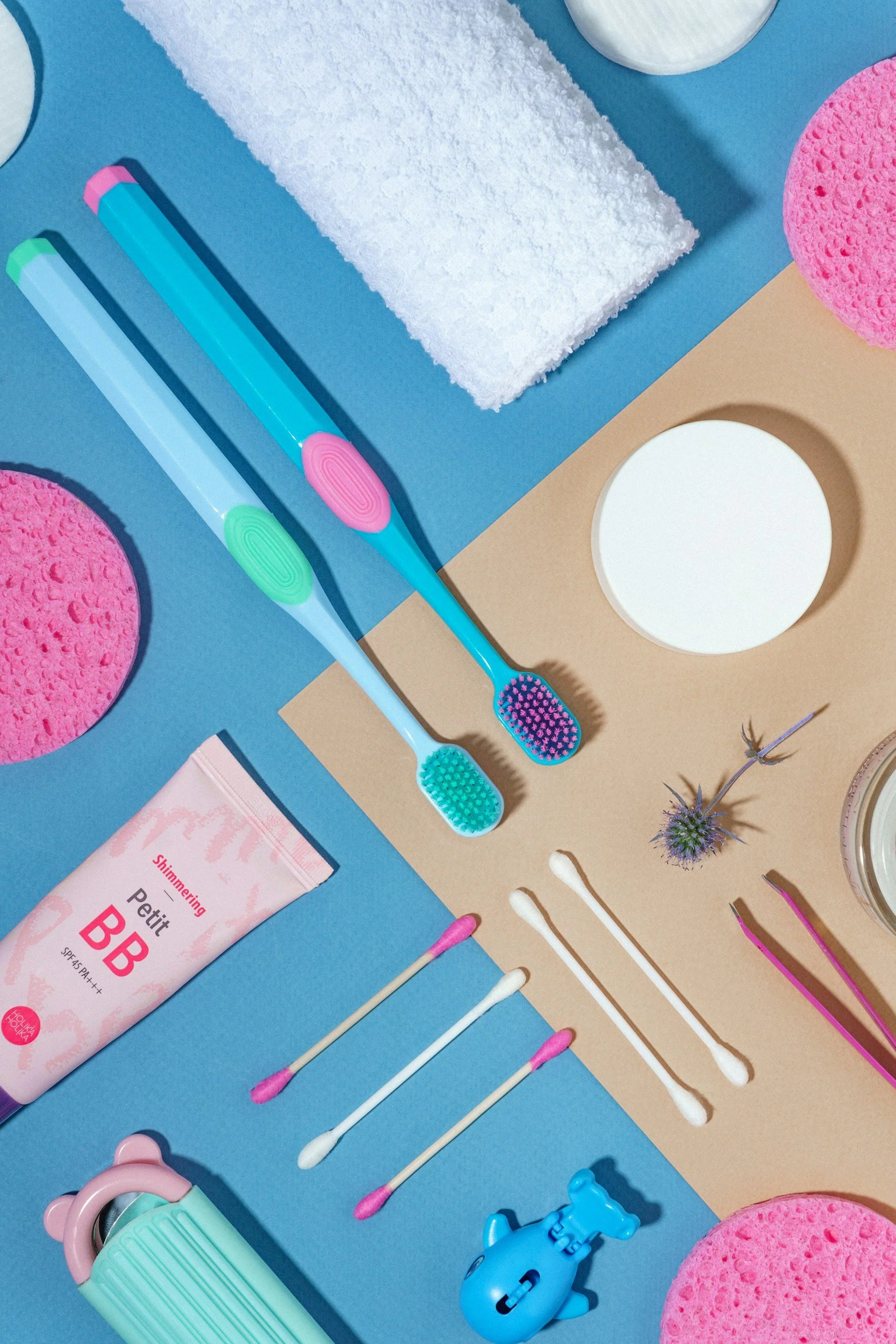 A flat lay of colorful bathroom and skincare items including two toothbrushes, cotton swabs, a tube of BB cream, a toy, sponges, a mirror, a flower, and a thermal water spray, arranged on blue and beige backgrounds.
