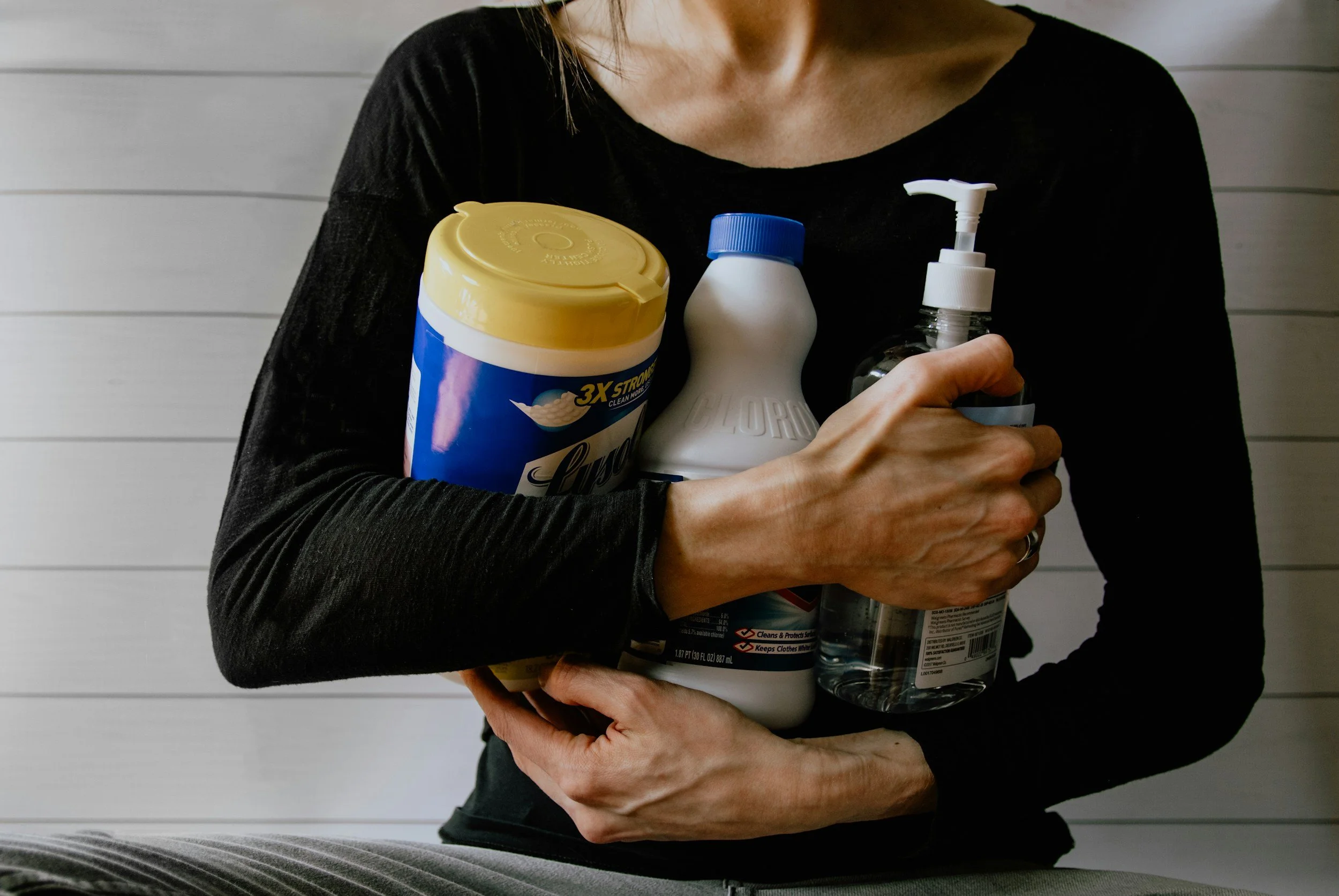 Person holding multiple household cleaning products, including a container of Lysol wipes, a bottle of bleach, and a spray bottle, against a white wooden wall background.