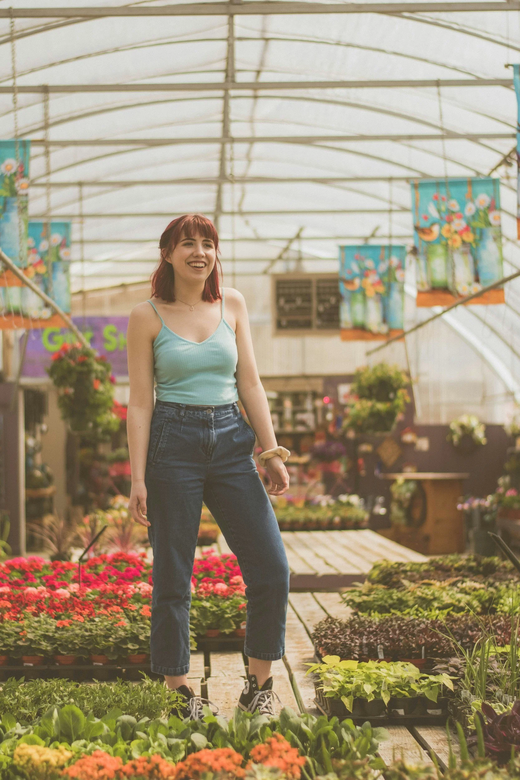A young woman with red hair wearing a light blue tank top, jeans, and sneakers, standing in a greenhouse with potted plants and flowers around her, smiling.