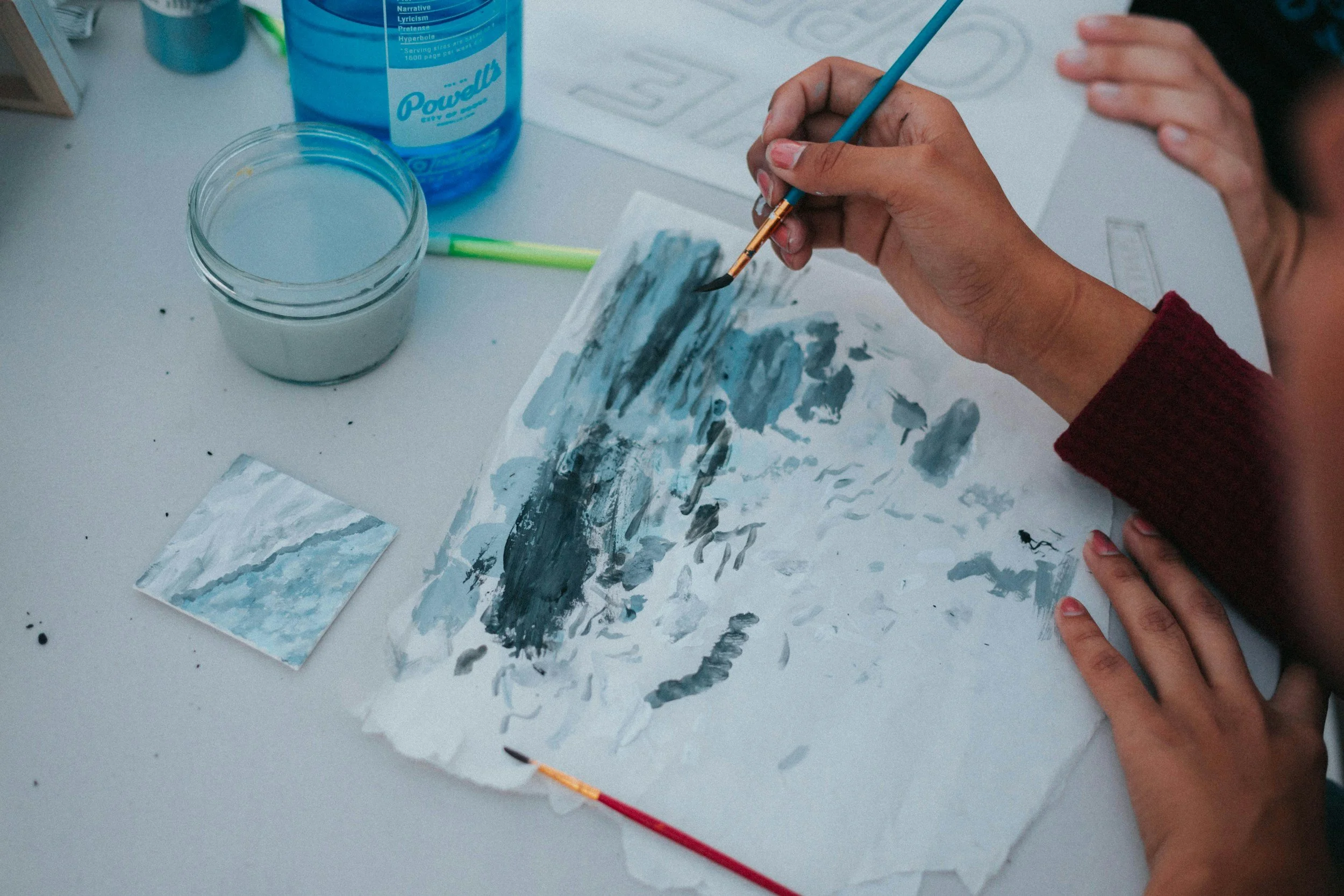 Person painting with blue, black, and gray watercolor paints on a sheet of paper, with art supplies and a small painted swatch nearby.