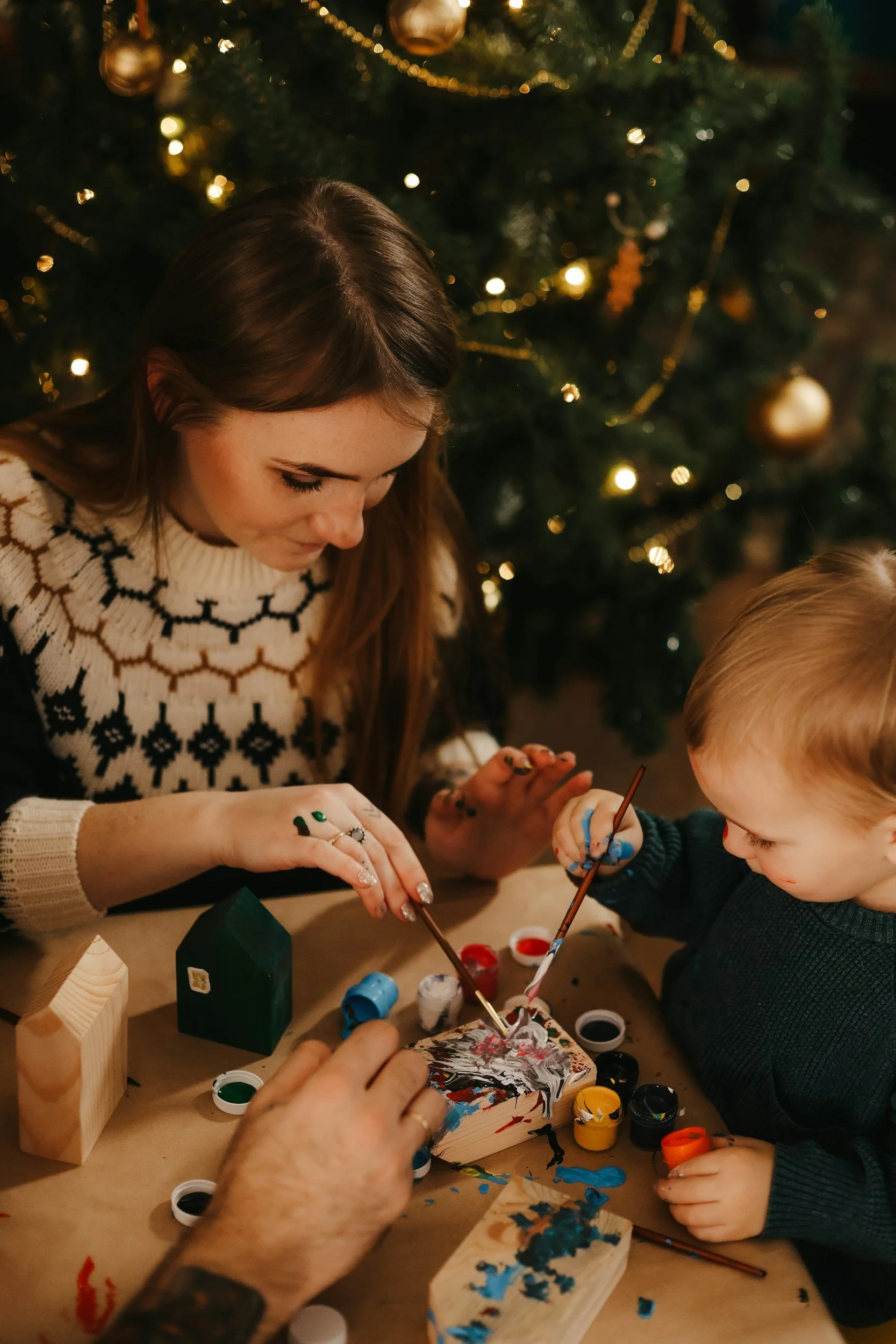 A woman and a young boy painting a small wooden house together at a Christmas-themed table, with a decorated Christmas tree in the background.