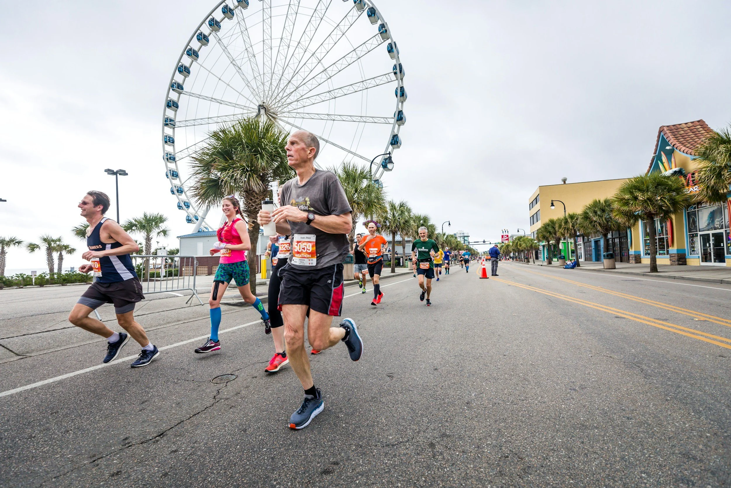 Marathon runners on a coastal street near a Ferris wheel, with palm trees and colorful buildings, under a cloudy sky.