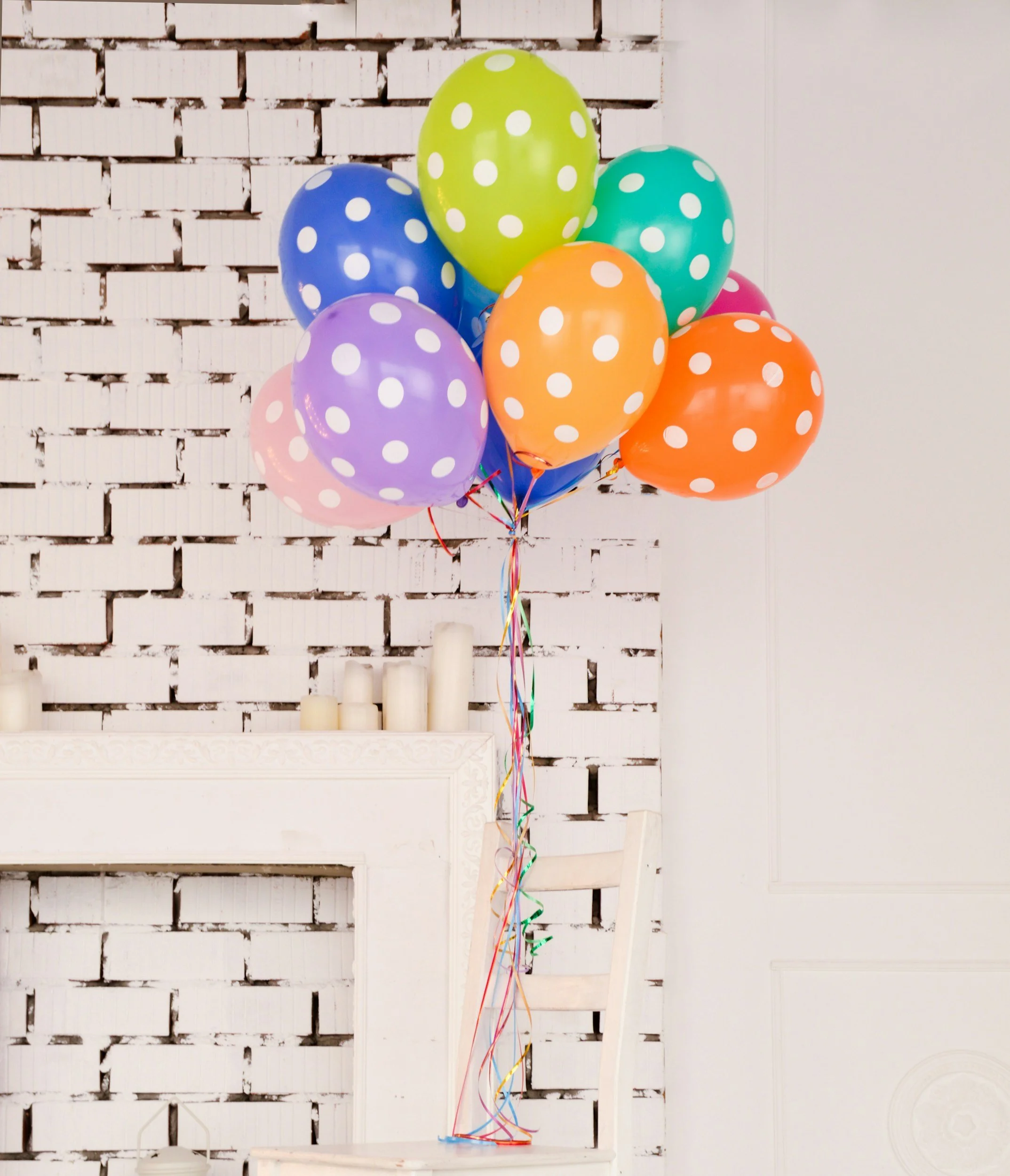 Colorful polka dot balloons tied together, floating in front of a white brick wall and white fireplace mantel.