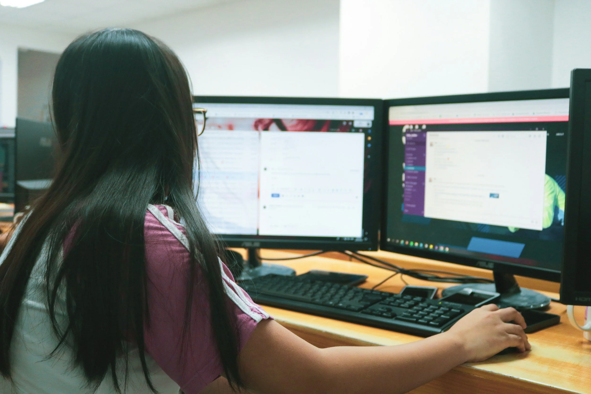 A woman with long dark hair and glasses working at a desk with two computer monitors.