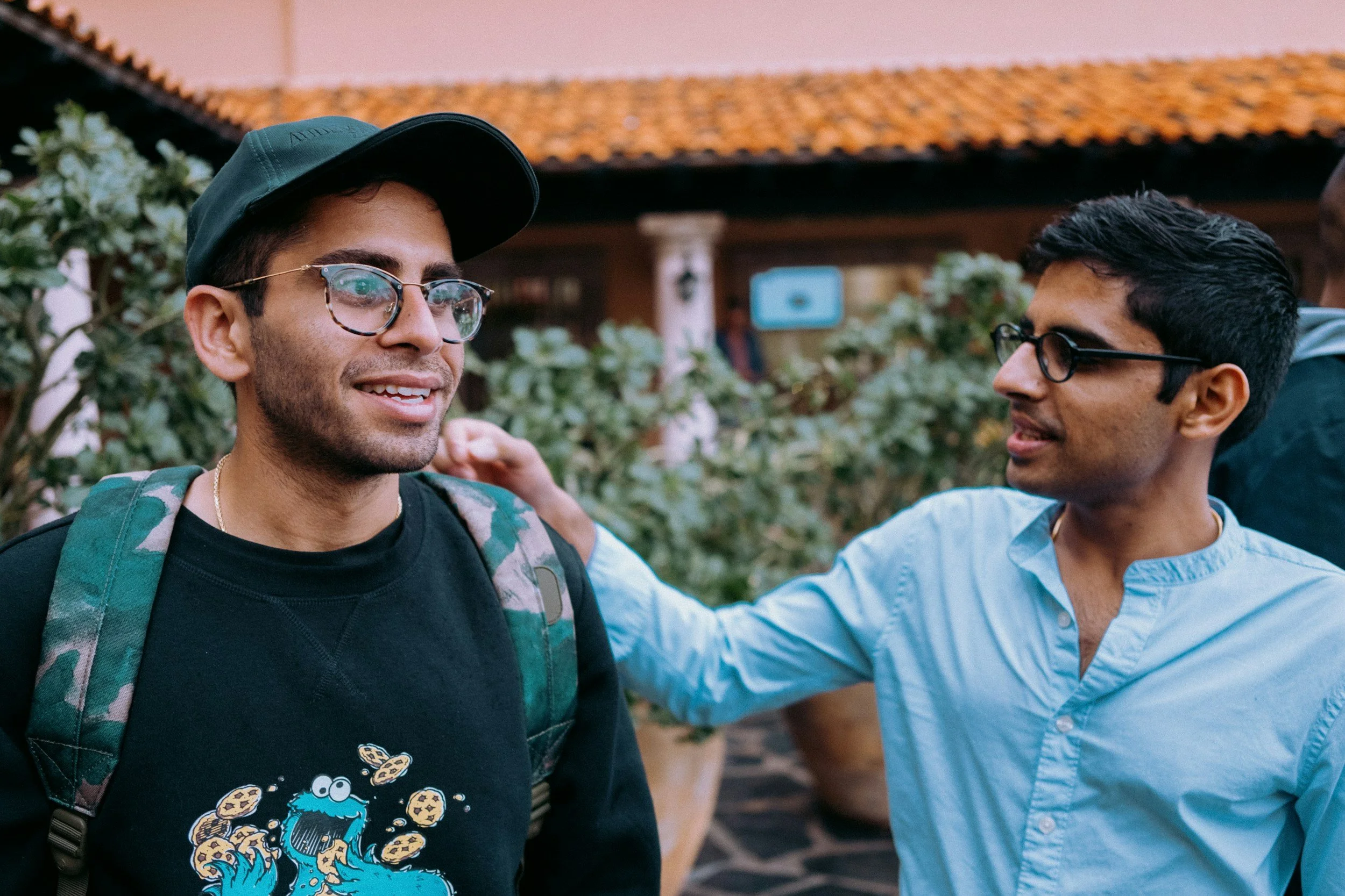 Two men, one wearing glasses, a black cap, and a backpack, smiling and talking to each other outdoors near plants and a building with a tiled roof.