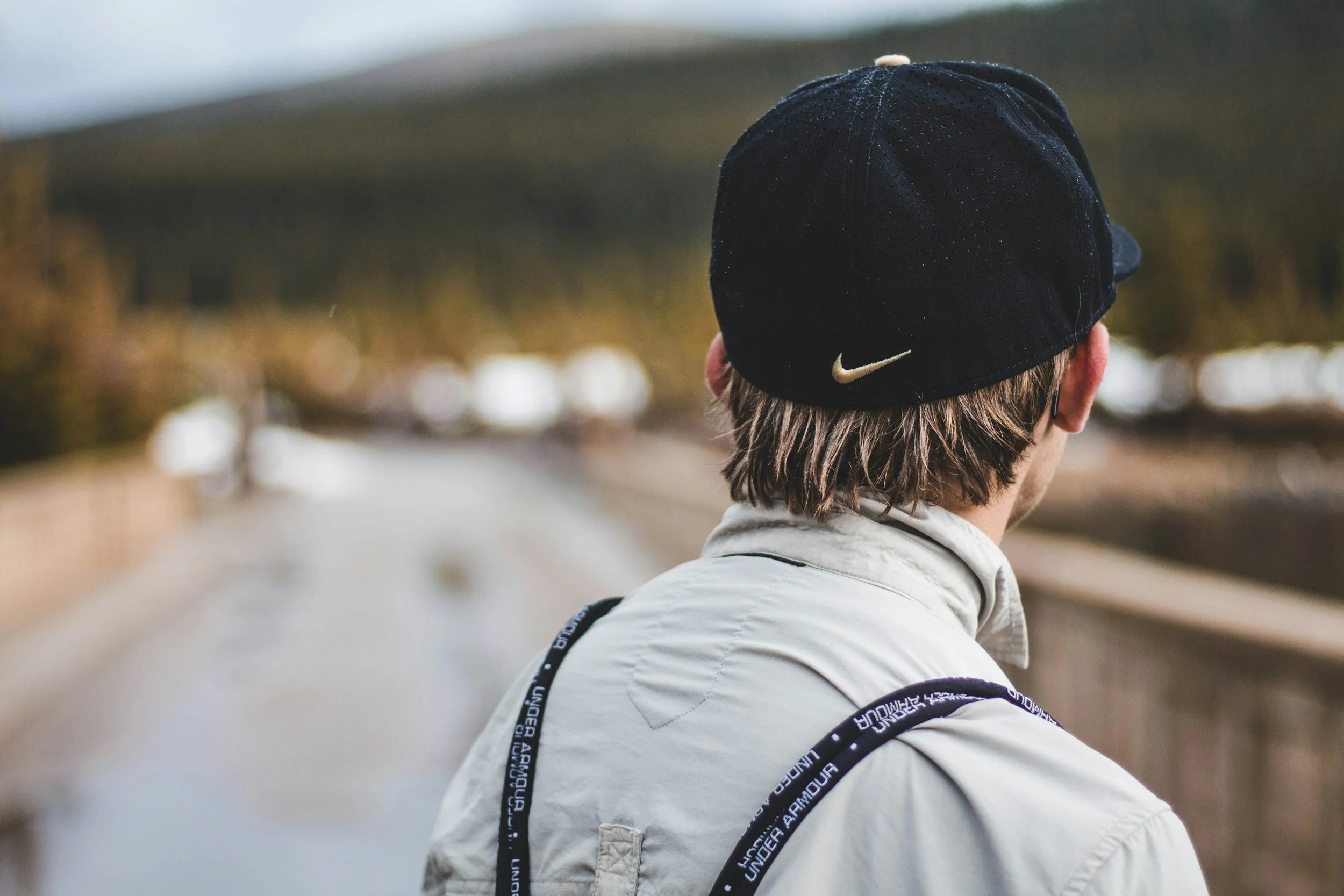 Person wearing a black Nike cap and white jacket with black shoulder straps, standing on a bridge, looking away at a blurred outdoor landscape with trees.