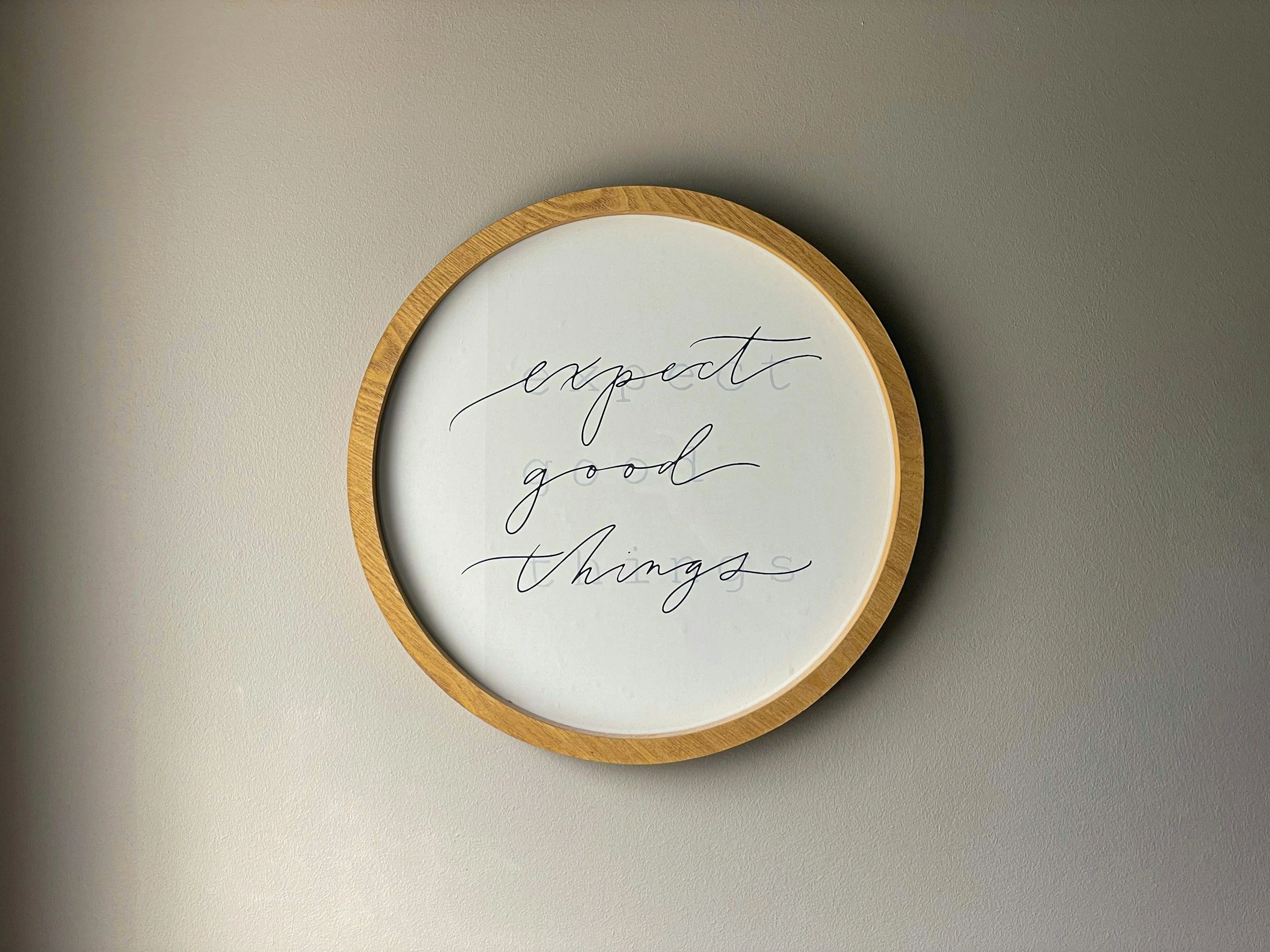 A round wall sign with a wooden frame displaying the words 'expect good things' written in cursive black font on a white background.