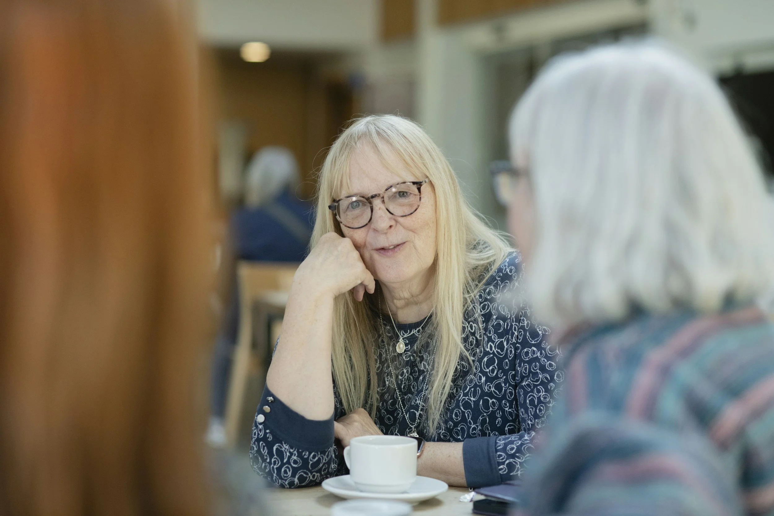 Two elderly women with light-colored hair and glasses are sitting at a table in a cafe or restaurant, engaged in conversation. One woman is resting her chin on her hand, with a cup of coffee in front of her.