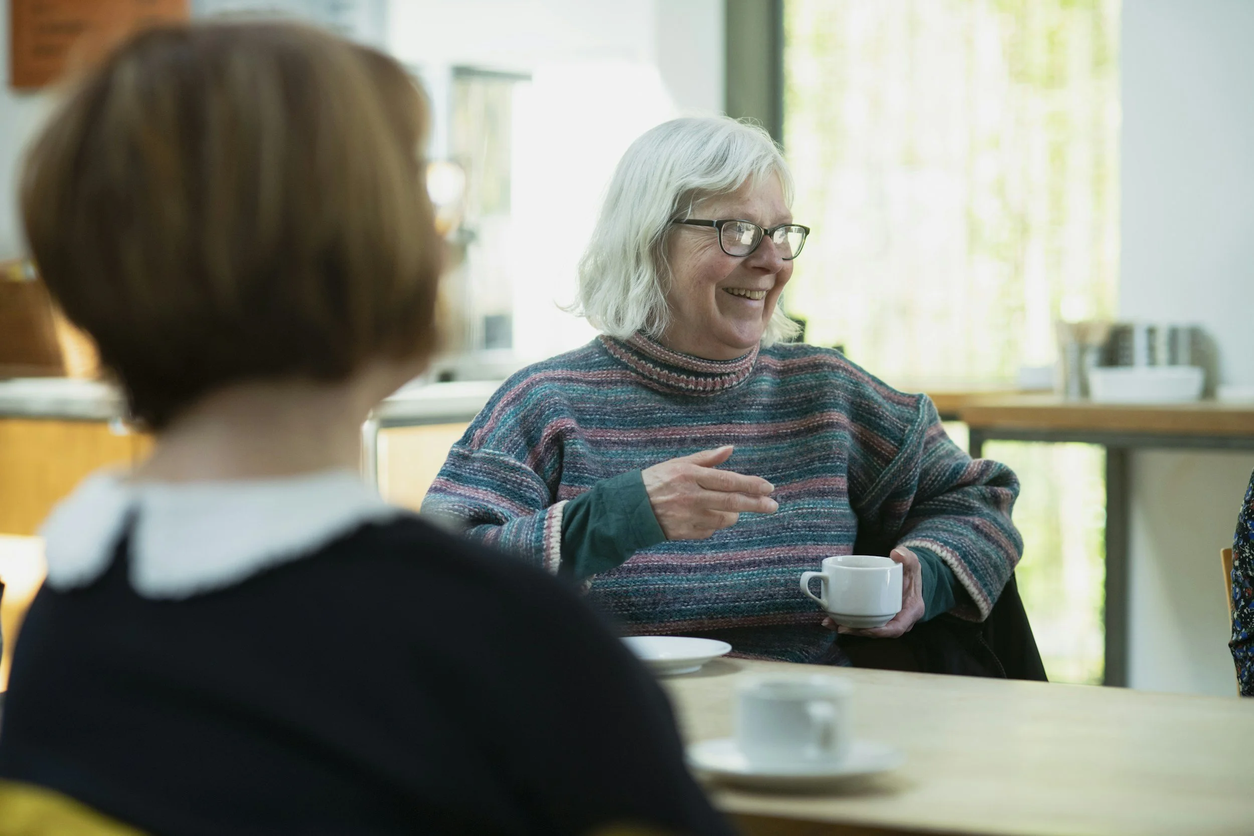 An elderly woman with glasses and a colorful sweater smiling and talking while holding a white coffee mug, sitting at a table with tea cups in a bright room with large windows.