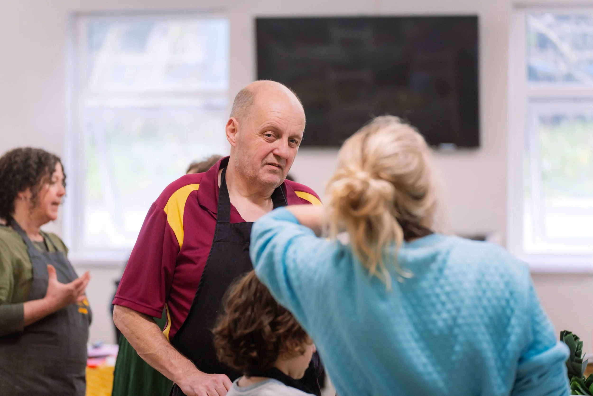 A man with a bald head and wearing a maroon shirt with yellow accents, and a black apron, standing and talking to a woman with blonde hair tied back, wearing a blue sweater. Other people are in the background, indoors with white walls and windows.