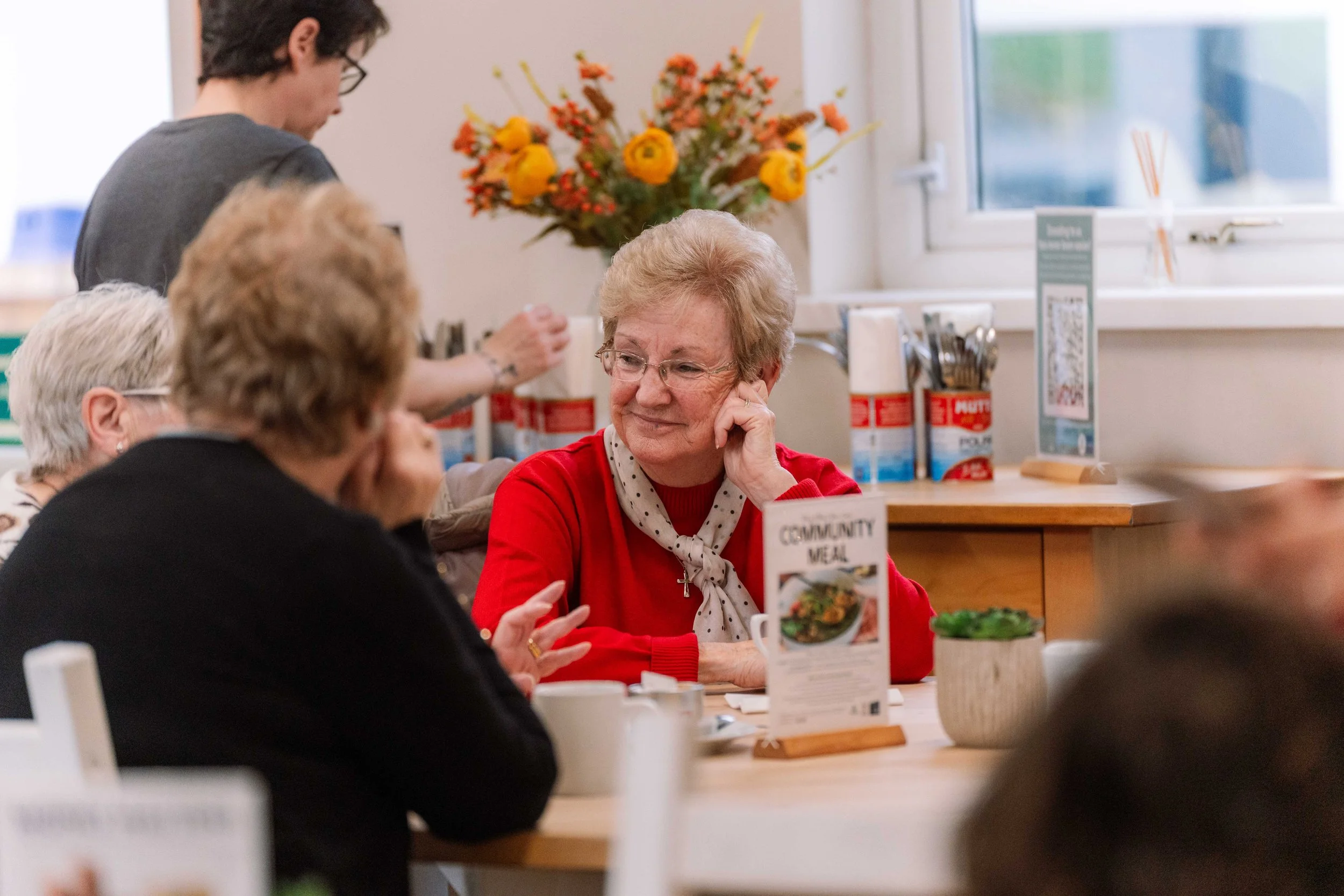 A group of elderly women sitting at a table in a community meal setting, with one woman wearing a red sweater and polka dot scarf, smiling and listening to others, with a server in the background and a bouquet of yellow and orange flowers on the window sill.