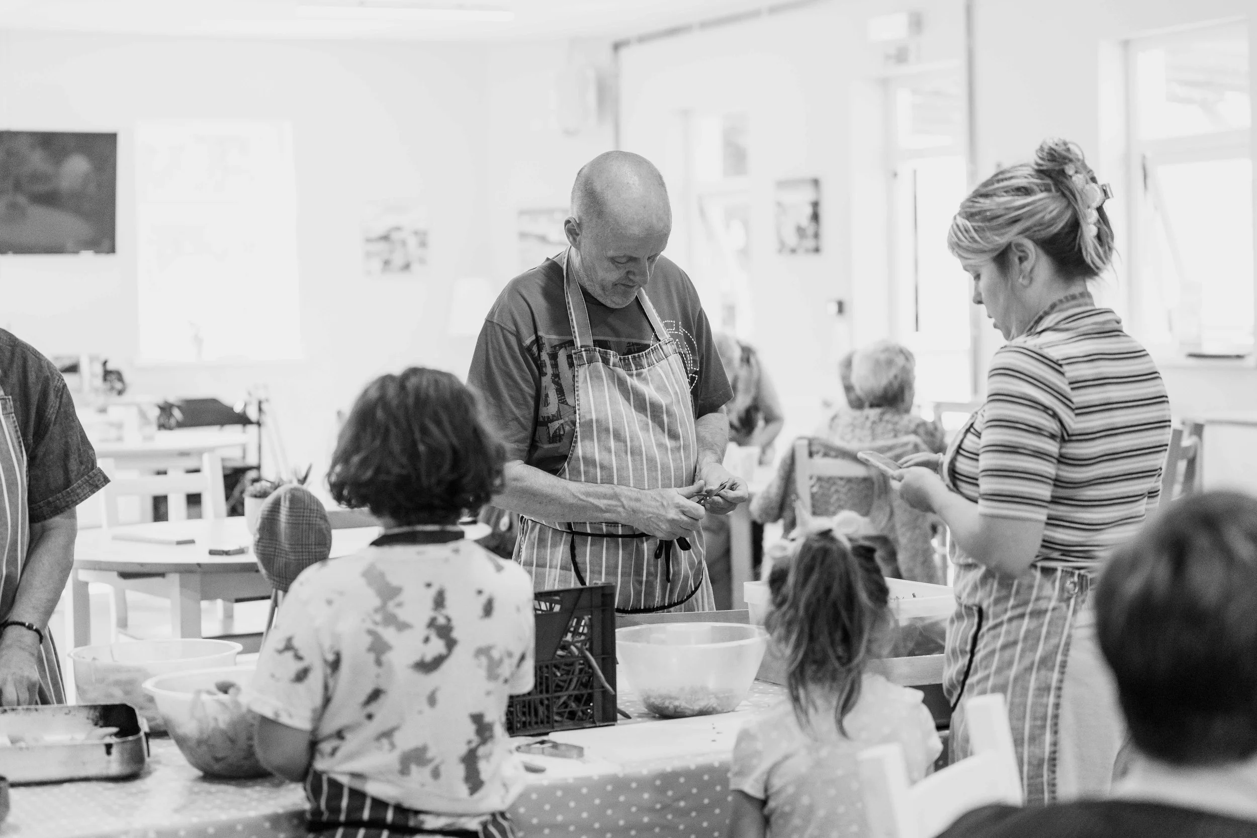People in a kitchen with aprons preparing food, with children and adults around.