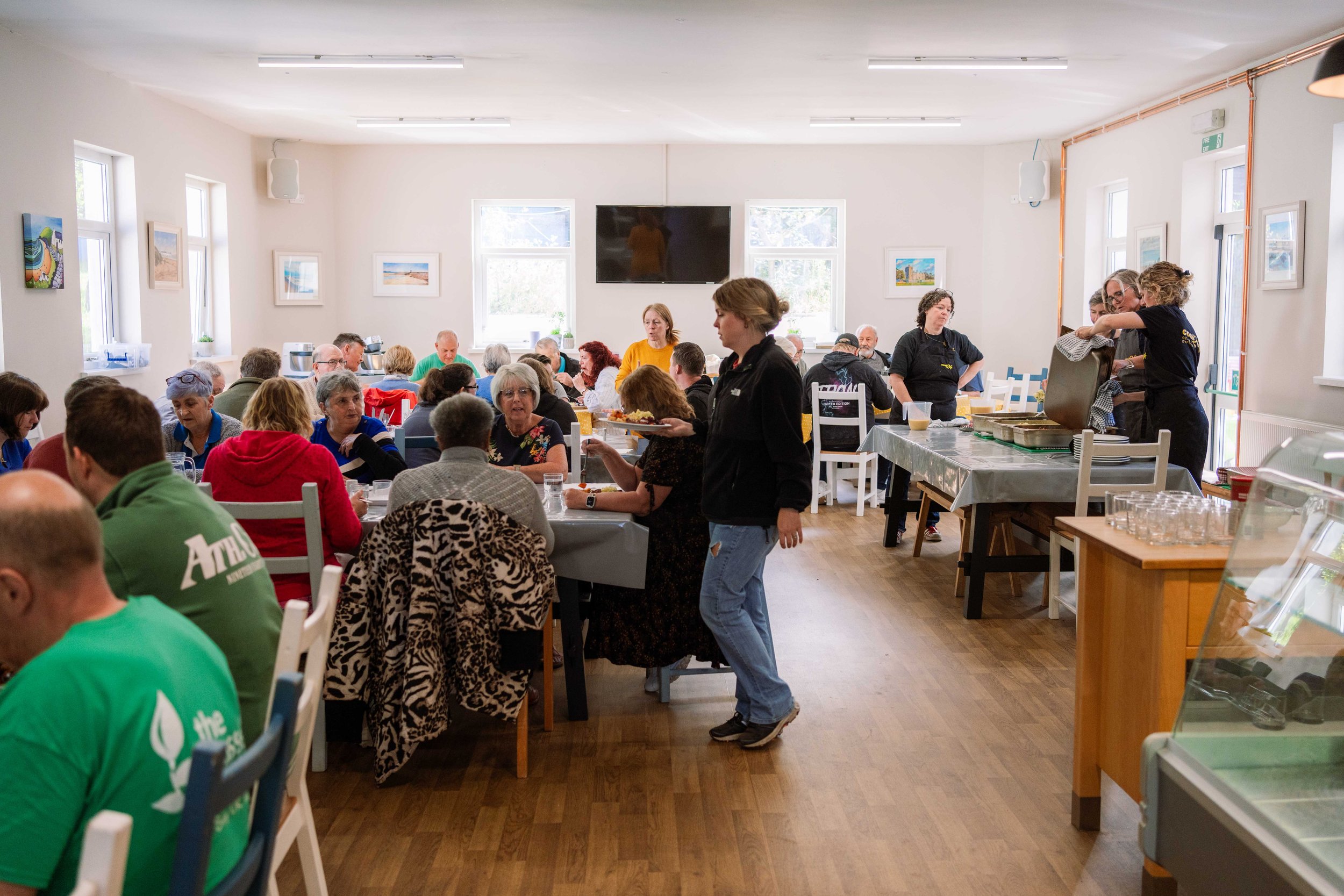 A busy communal dining room filled with people eating and talking, with a buffet table on the right where staff serve food.