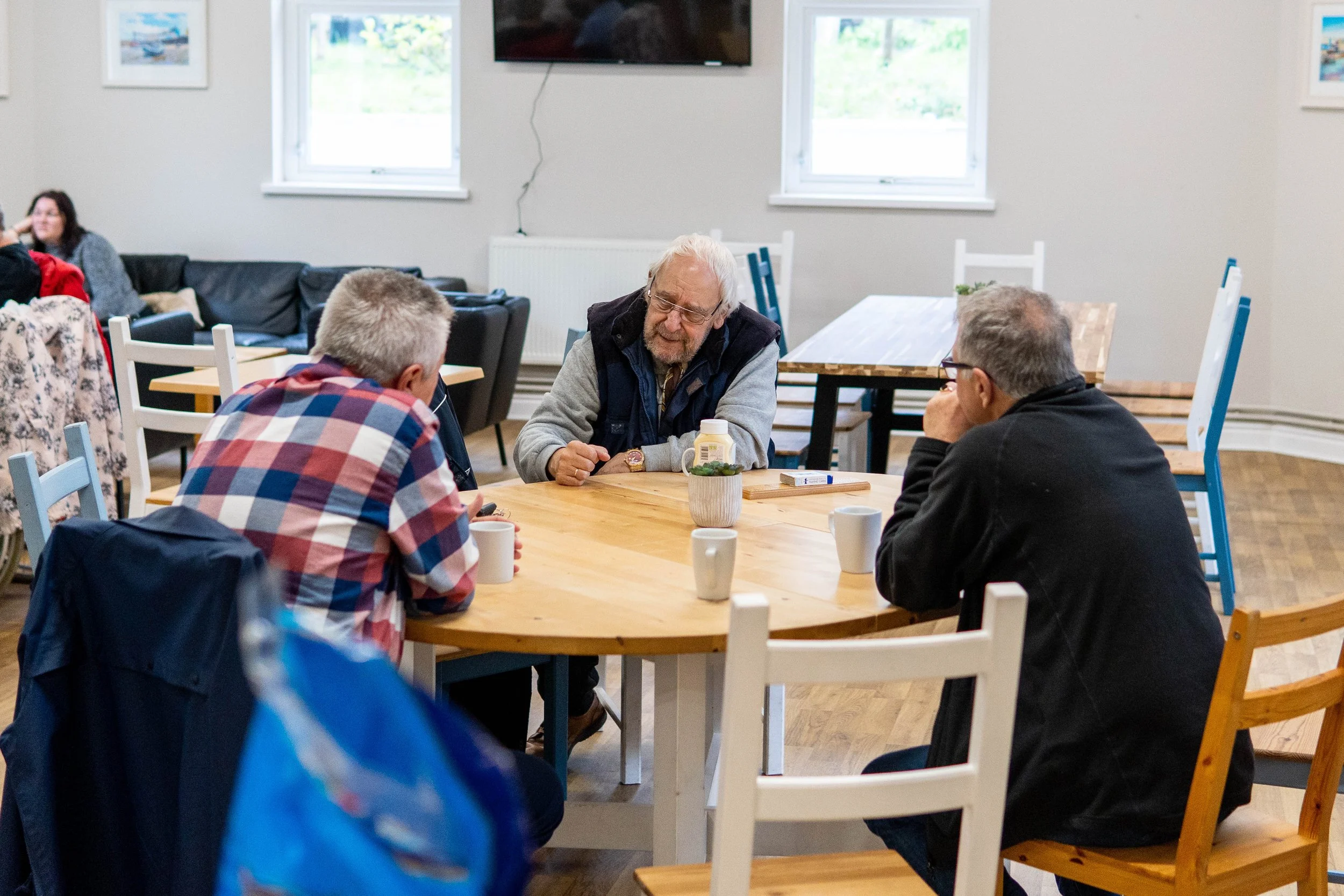 Three men sitting around a round wooden table having a conversation in a bright room with white walls and windows, with another person visible in the background.