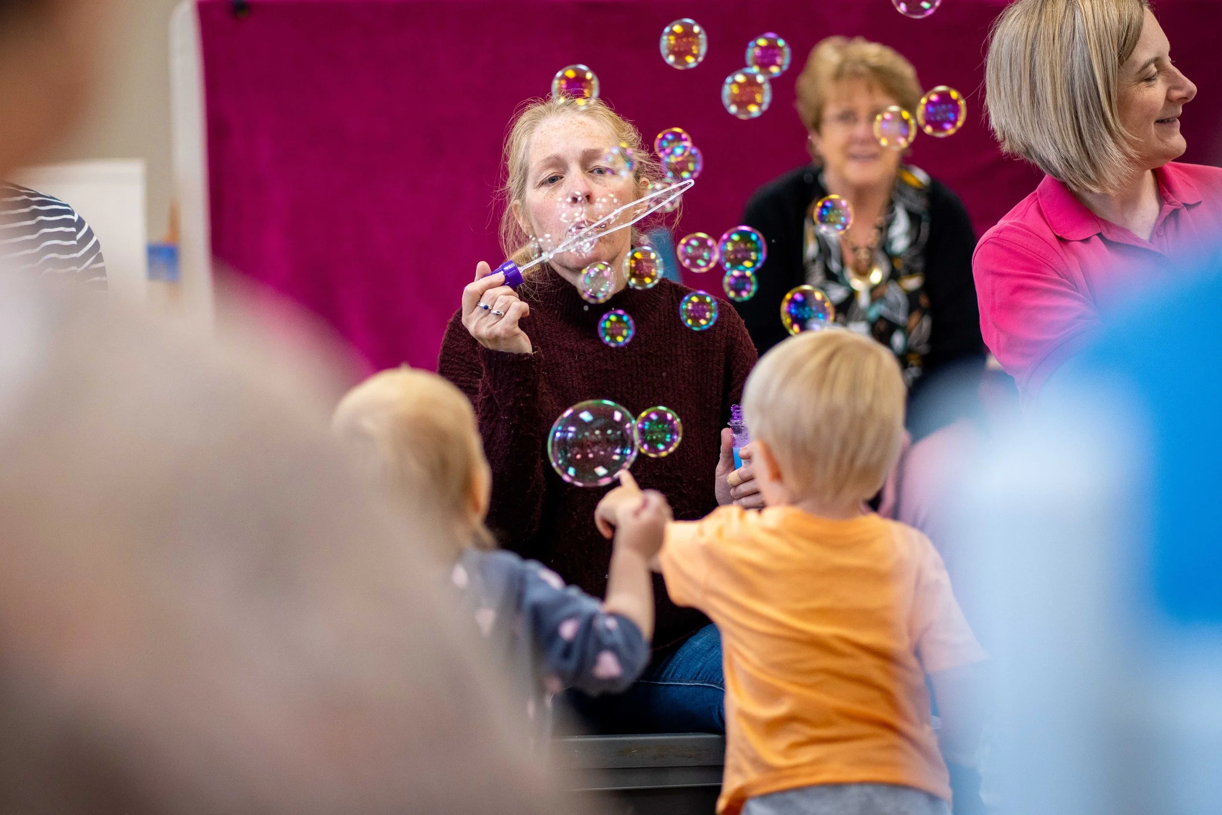 A woman blowing bubbles at a children's party with adults and children participating, with a maroon backdrop and pink wall.