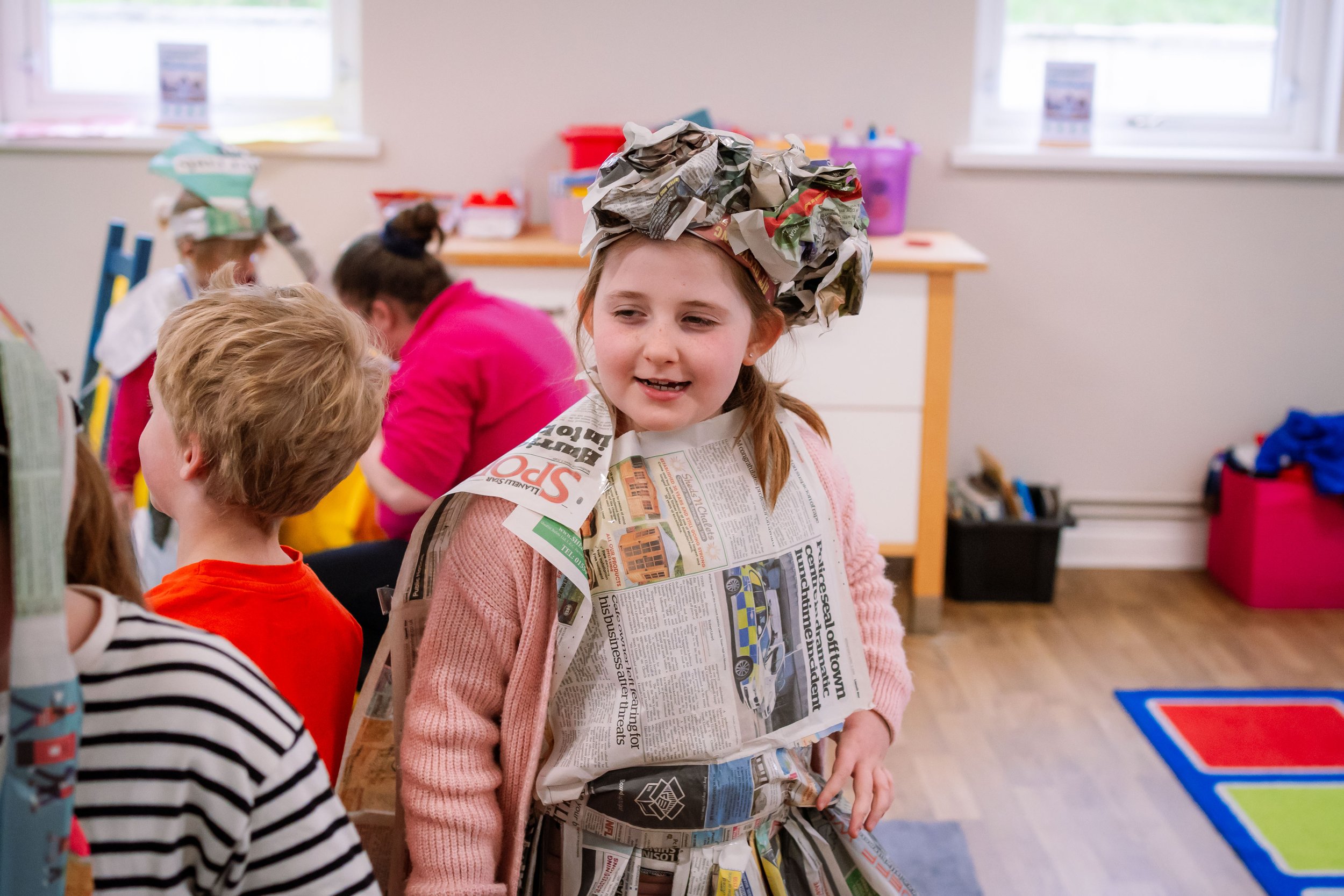 A young girl in a pink sweater and newspaper costume, wearing a newspaper hat, standing among other children in a classroom.