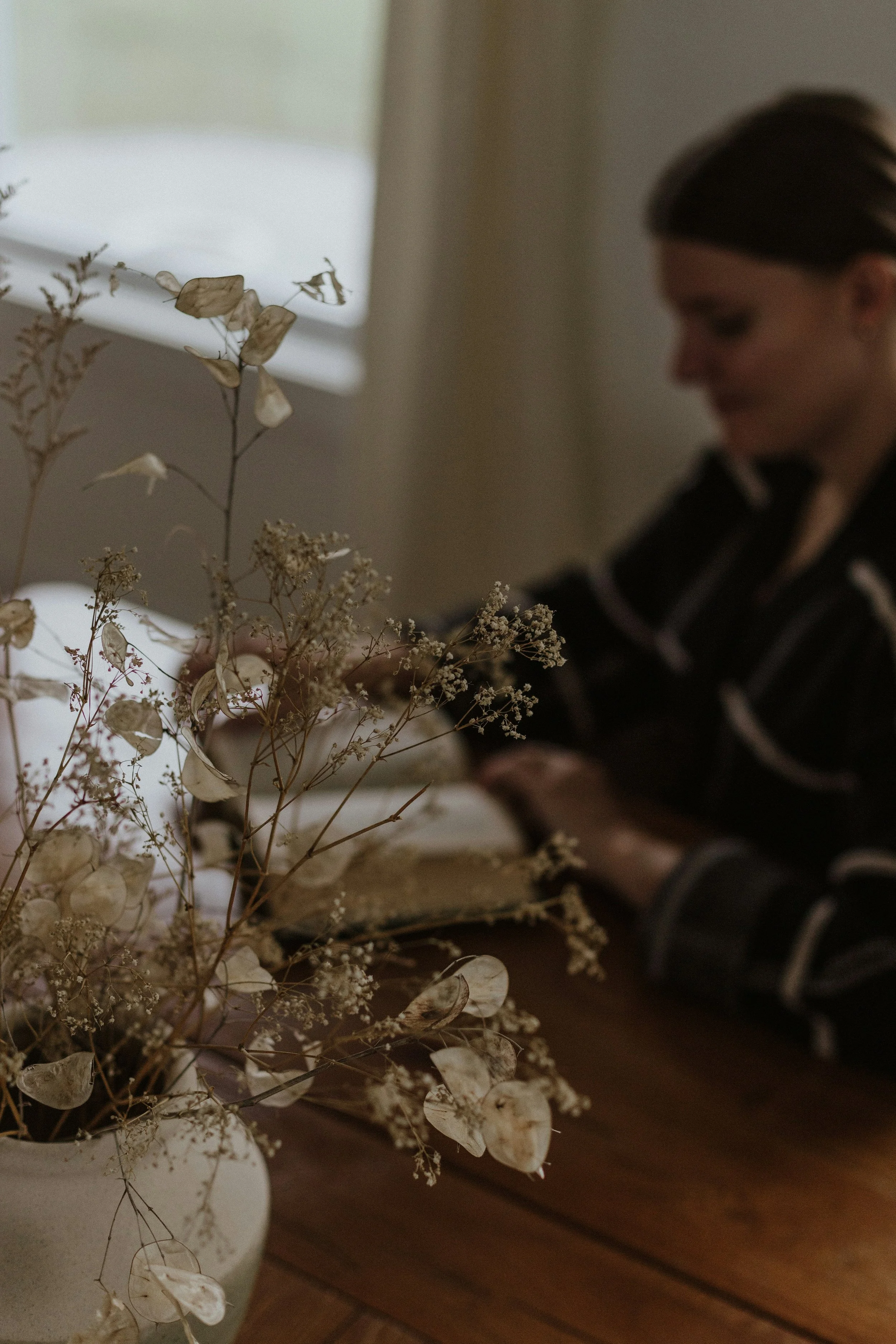 A woman in a dark striped shirt arranging or working with dried flowers in a vase on a wooden table, with a window in the background.
